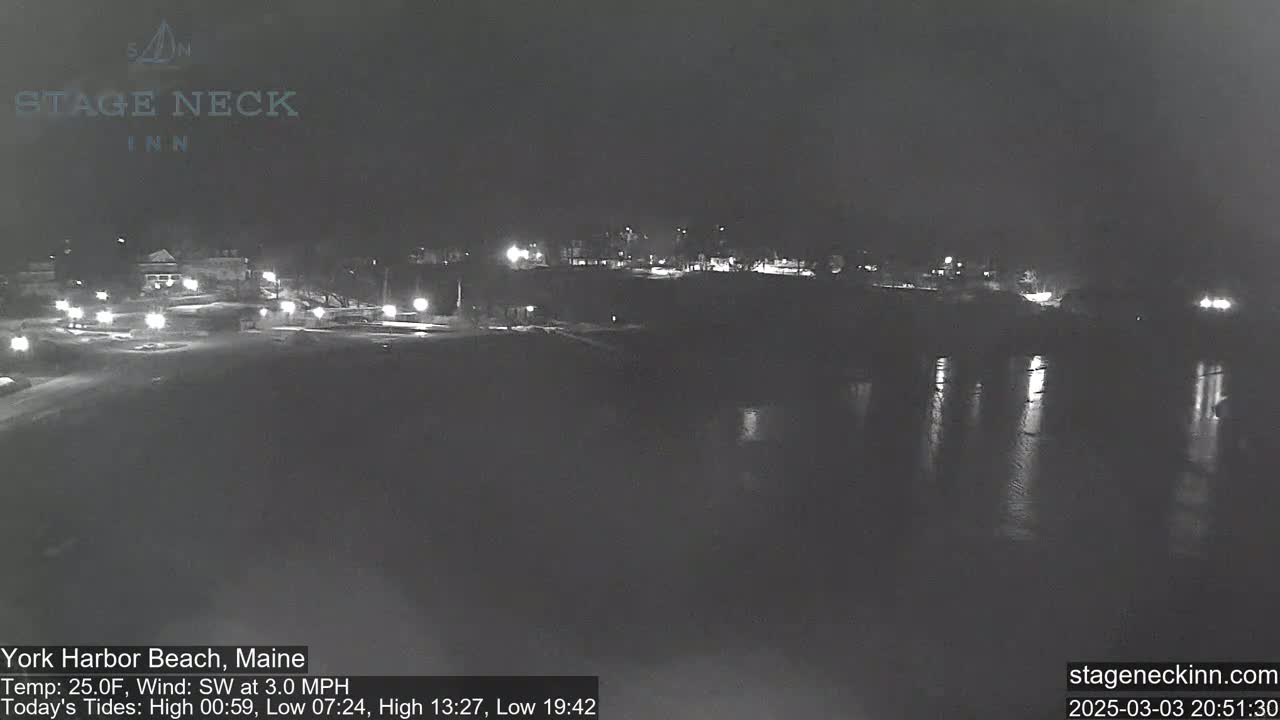 A nighttime view of a dark, calm body of water reflecting lights from a shoreline with buildings and sparse streetlights.