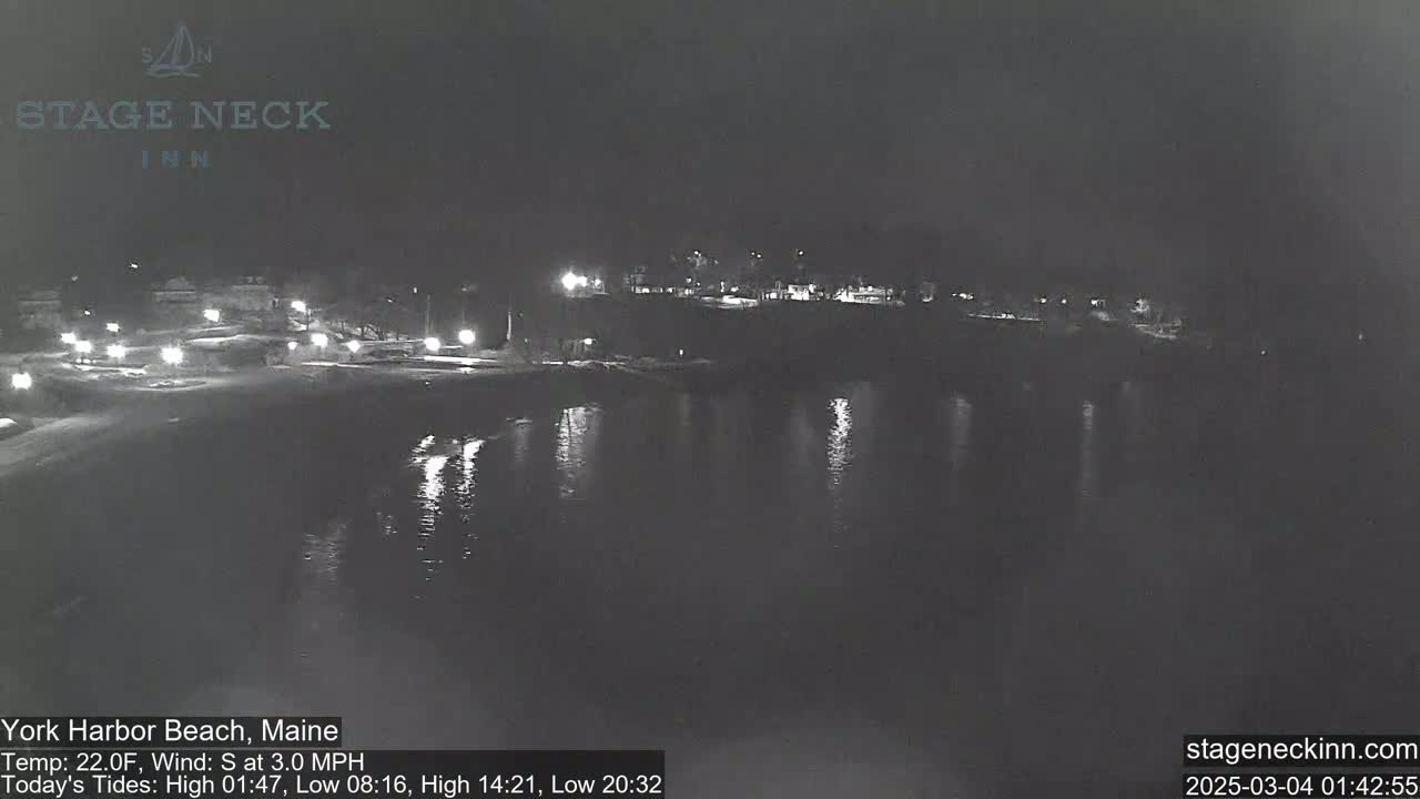 A nighttime view of a dark, calm body of water reflecting lights from a shoreline with buildings and sparsely lit walkways.
