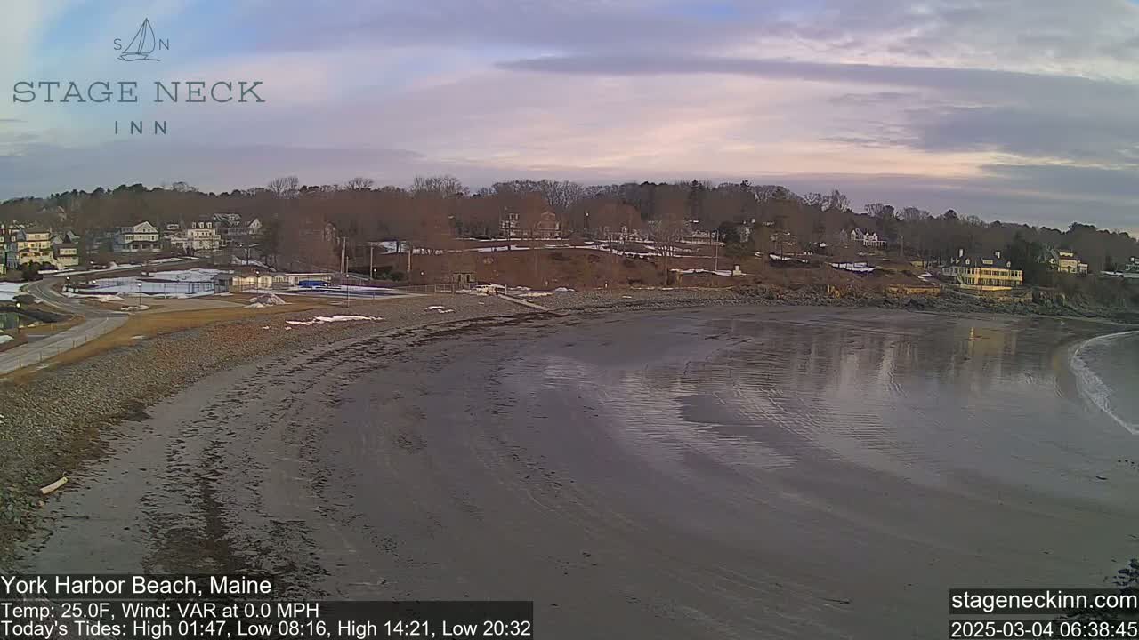 A mostly calm, sandy beach at low tide stretches out to a calm body of water, under a partly cloudy, dusky sky, with buildings and trees visible in the background.