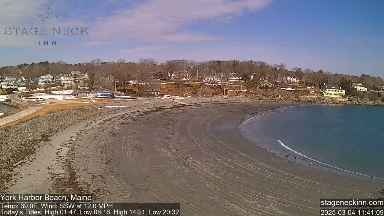 A sandy beach curves along a calm bay under a clear blue sky, with buildings and trees visible on the elevated land behind it.