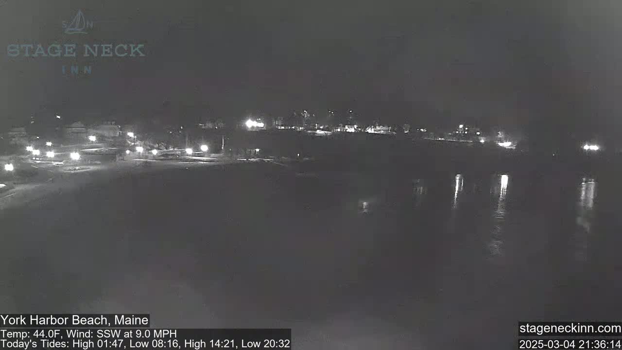 A nighttime, low-light view of a calm body of water reflecting a dimly lit shoreline with buildings and sparse vegetation.