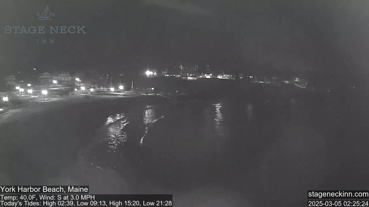 A nighttime view of a dark beach and calm water, with a few lights visible in the distance.