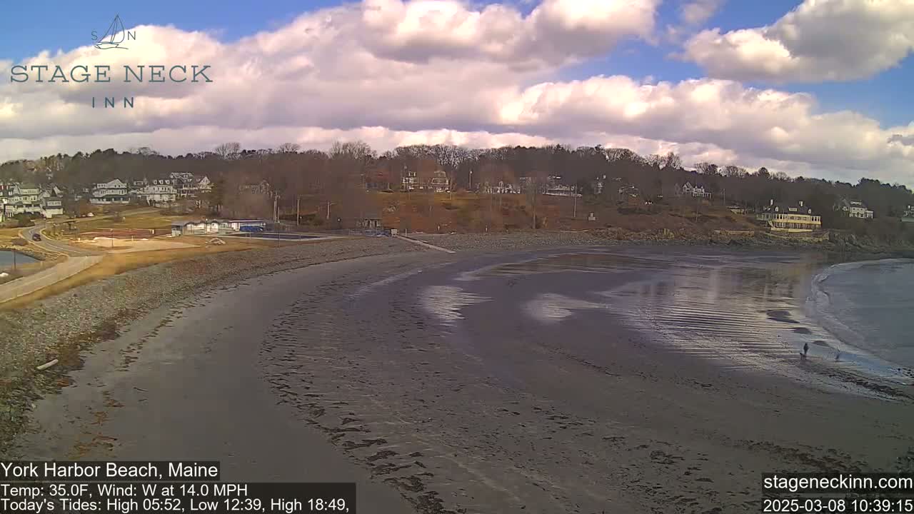 A mostly cloudy day shows a sandy beach with a calm ocean and a shoreline with buildings and trees.