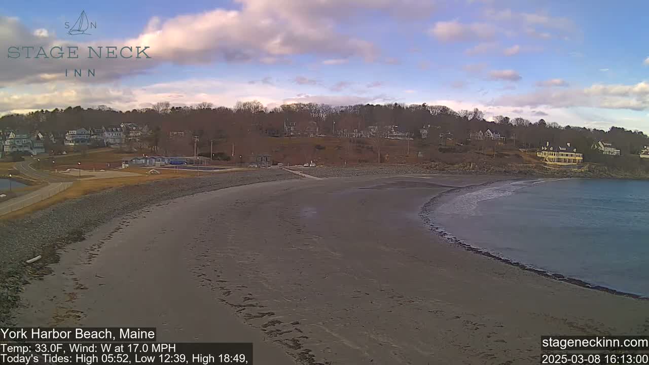 A sandy beach curves gently into a calm bay under a partly cloudy sky, with buildings and trees visible in the background.