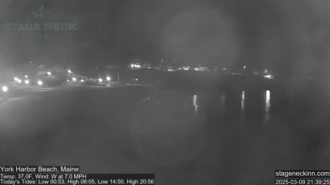 A nighttime, low-light view of a calm body of water reflecting lights from a nearby shore with buildings faintly visible in the distance under a dark, possibly overcast sky.