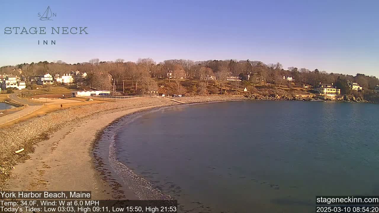 A sandy beach curves along a calm body of water, bordered by houses on a hill under a clear blue sky.