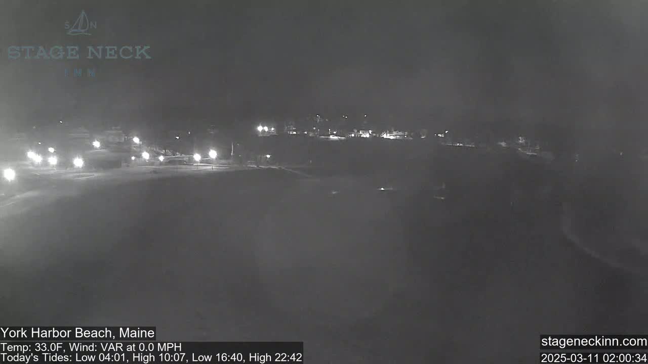 A nighttime, snowy scene shows a dimly lit road with sparse traffic and buildings in the distance under a dark sky.