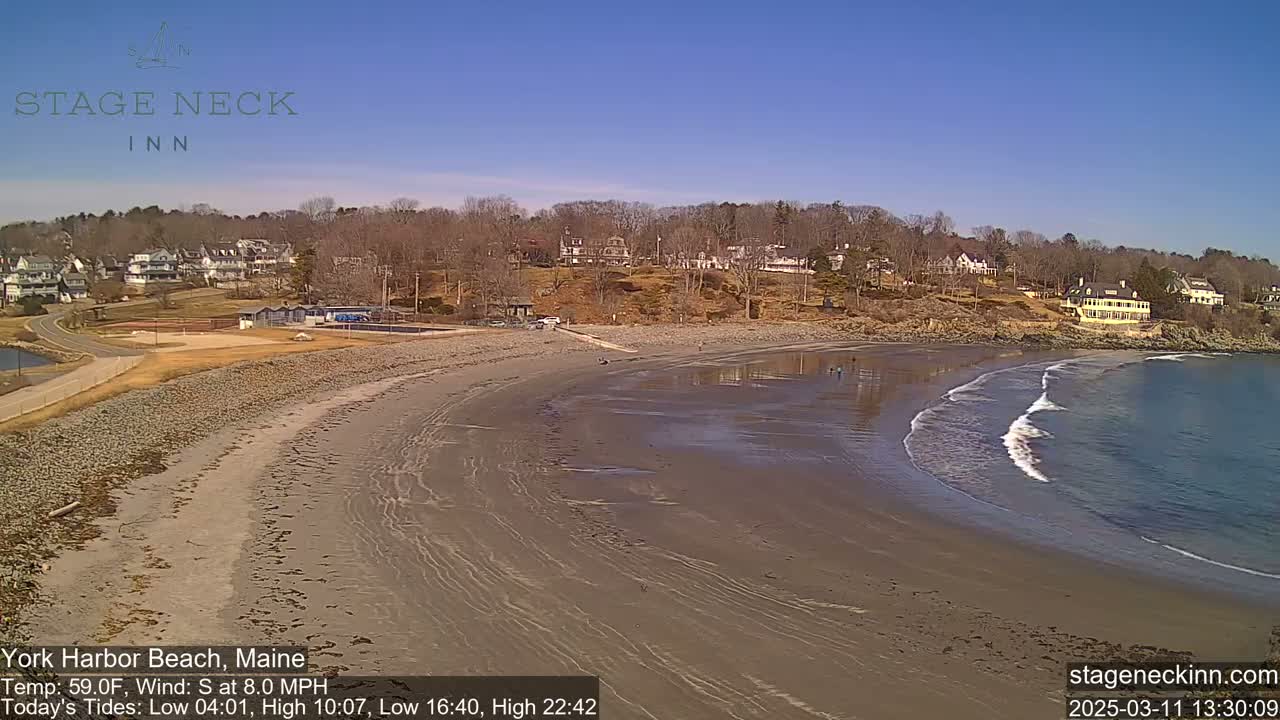 A sandy beach with gentle waves laps the shore under a clear blue sky, with buildings and trees visible in the background.