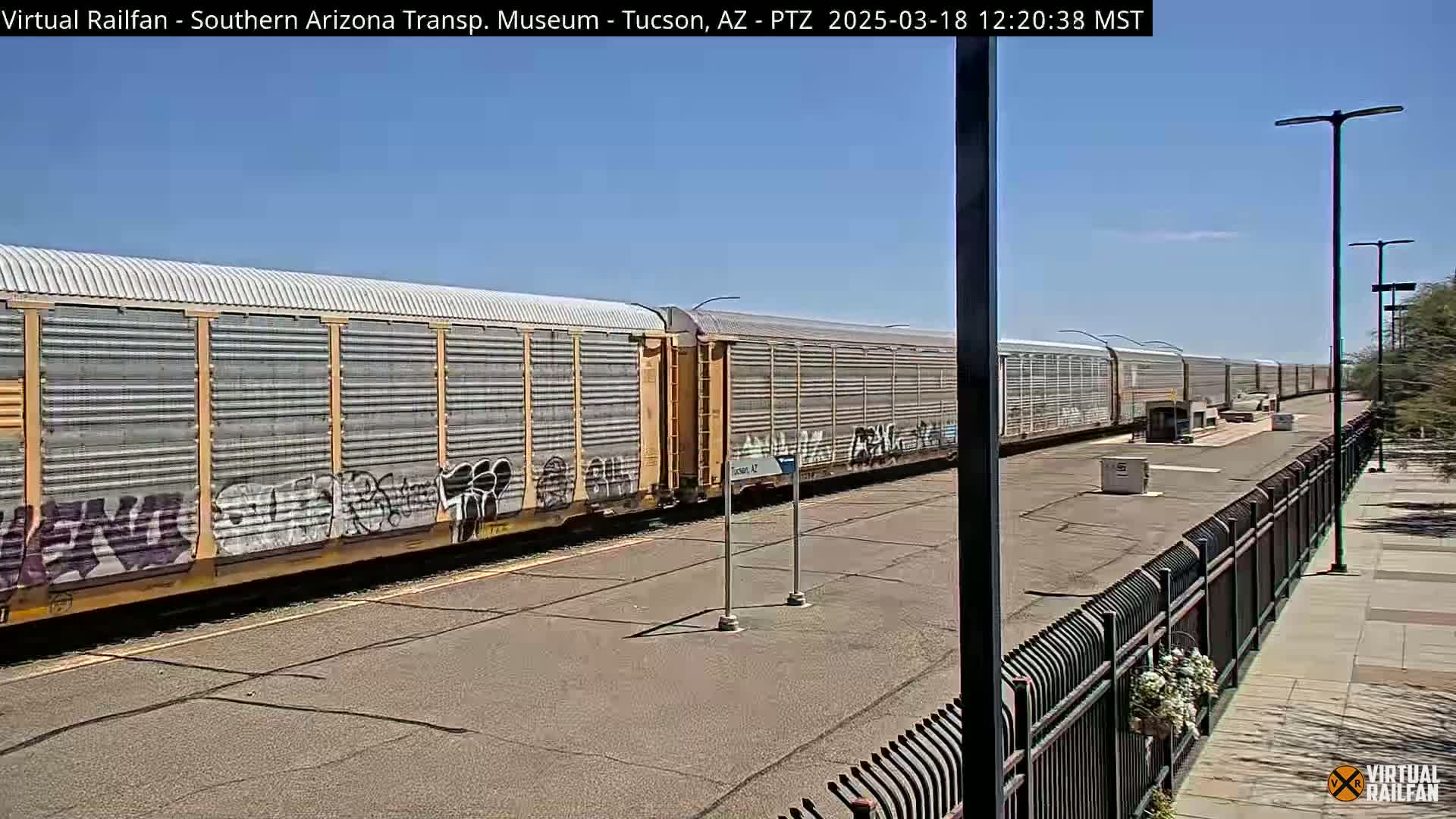 A long freight train with graffiti on its cars sits in a sunny, outdoor train yard next to a black metal fence.