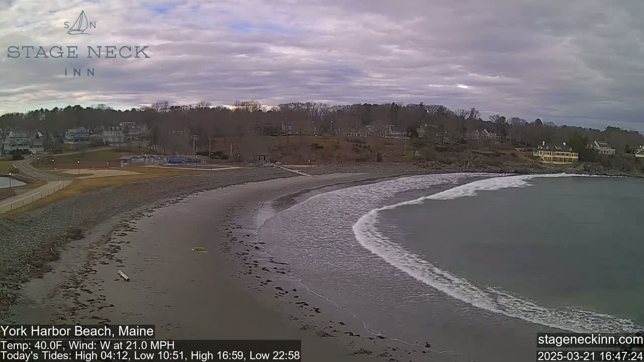 A sandy beach curves into a gray ocean with small waves under a cloudy sky.