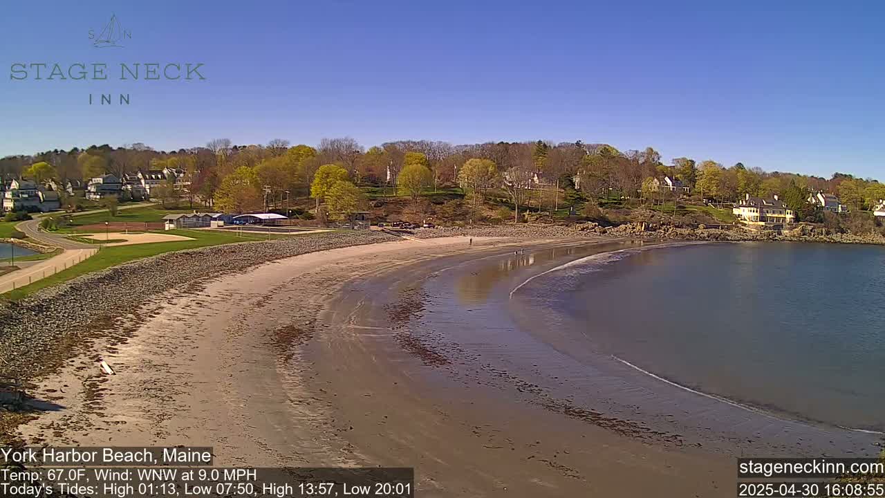 A sandy beach curves into calm water under a clear blue sky, with houses and trees visible on the hillside behind it.