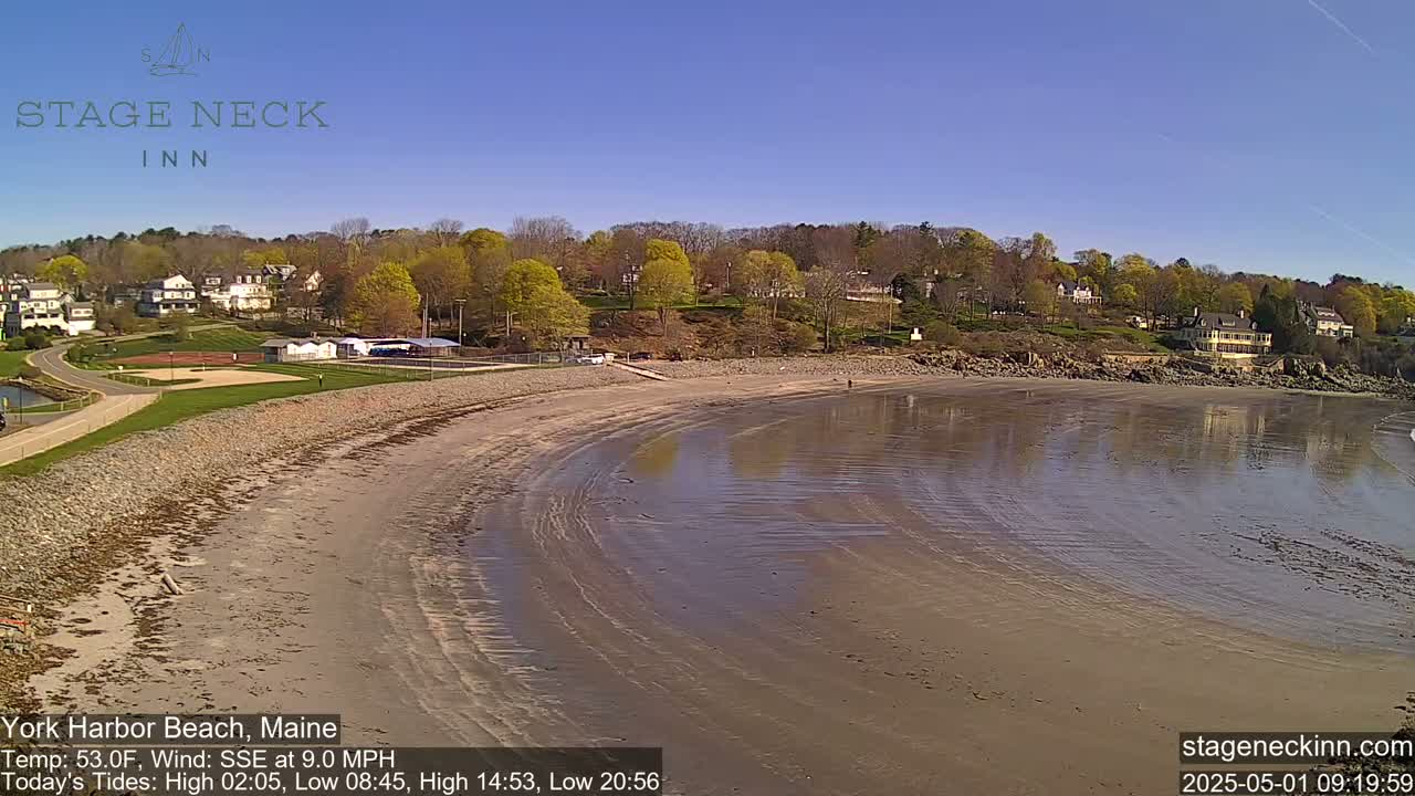A sandy beach curves around a calm body of water under a clear blue sky, with buildings and trees visible in the background.
