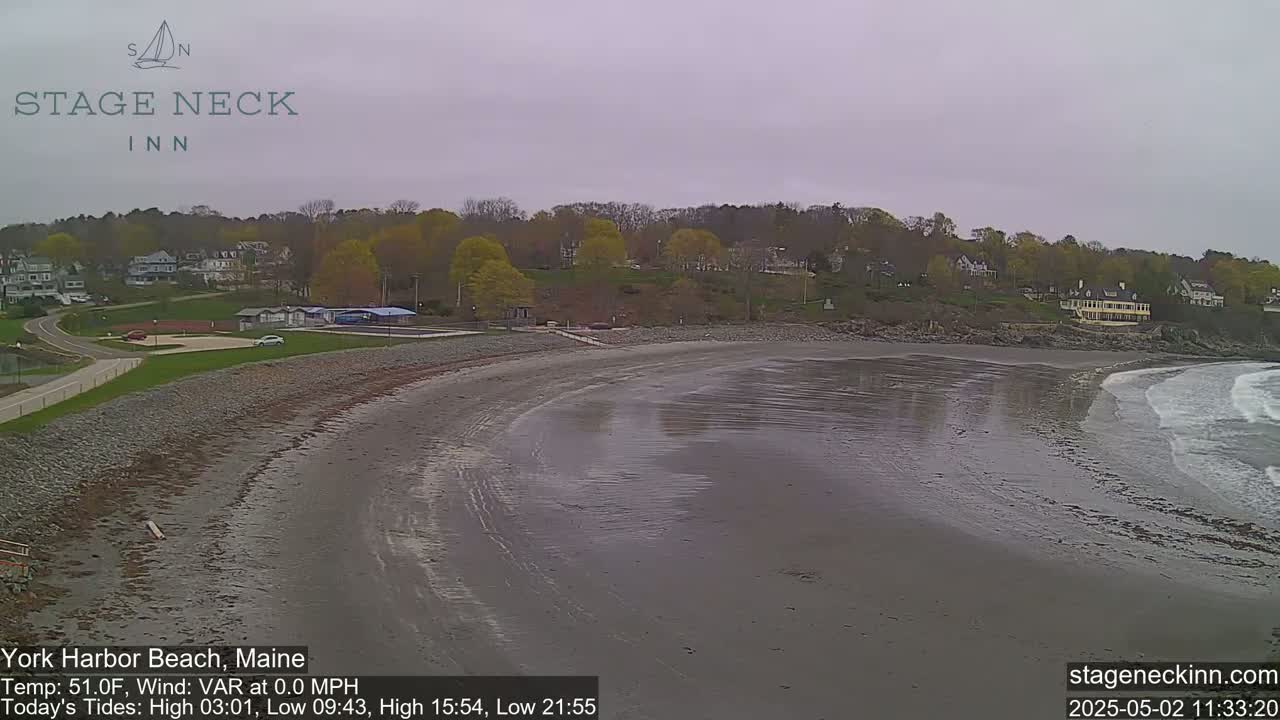 A mostly overcast day reveals a sandy beach curving gently into the ocean, with calm waves lapping the shore, backed by a line of trees and buildings.