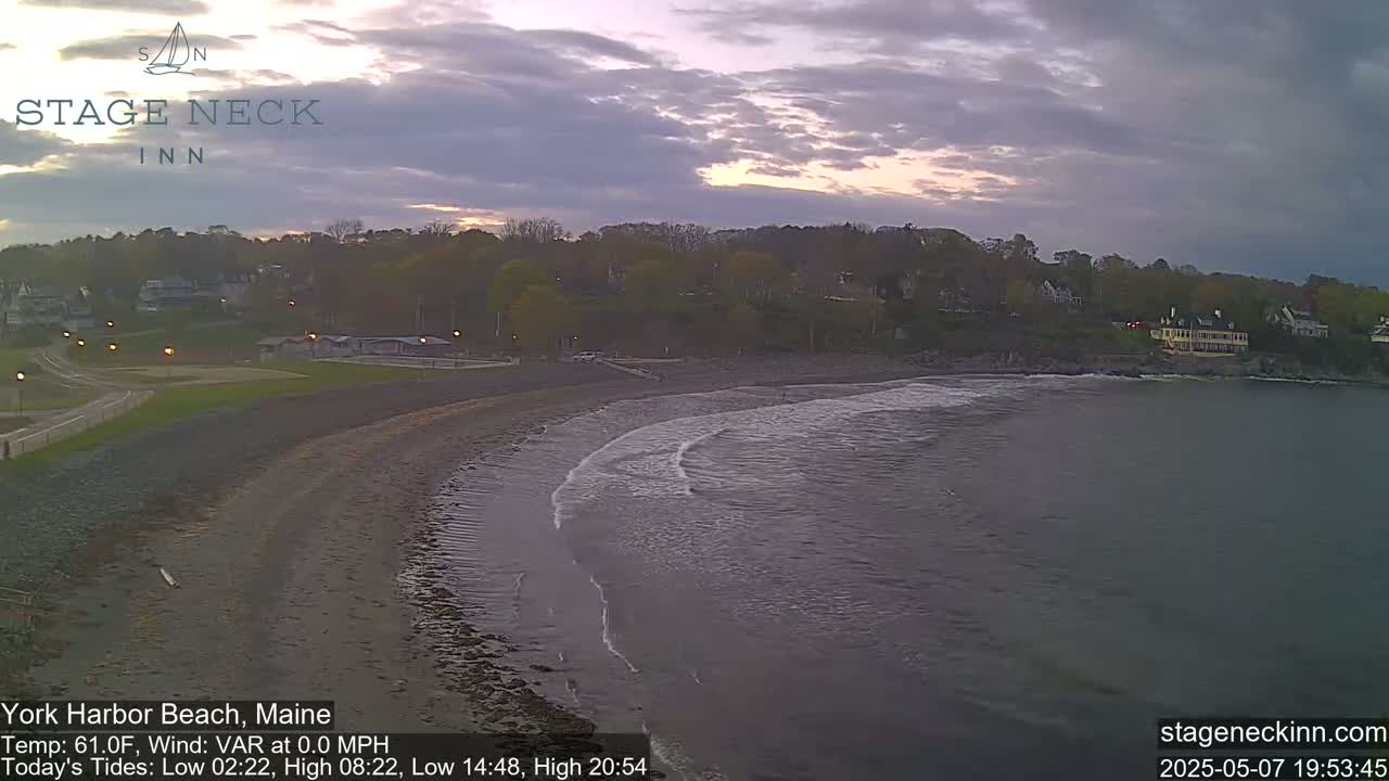 A sandy beach curves into calm, dark water under a partly cloudy, dusky sky.