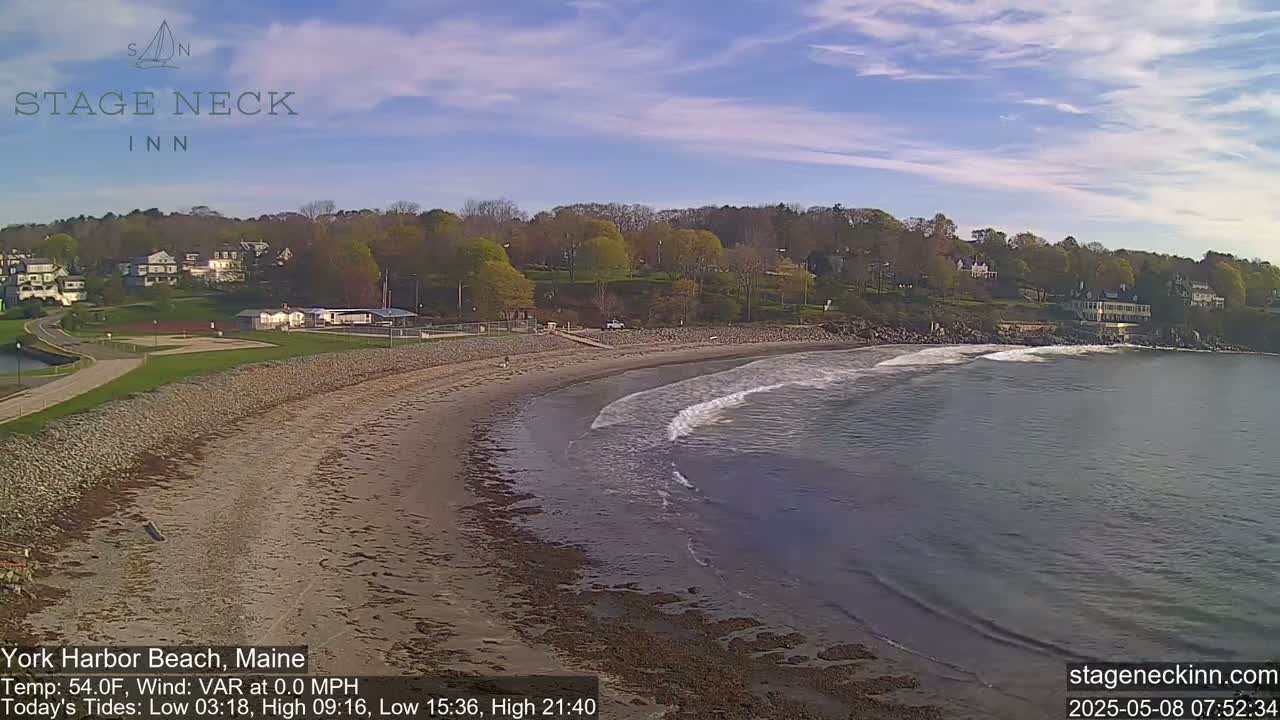 A sandy beach curves gently into calm ocean water under a mostly clear sky, with green trees and buildings visible in the background.
