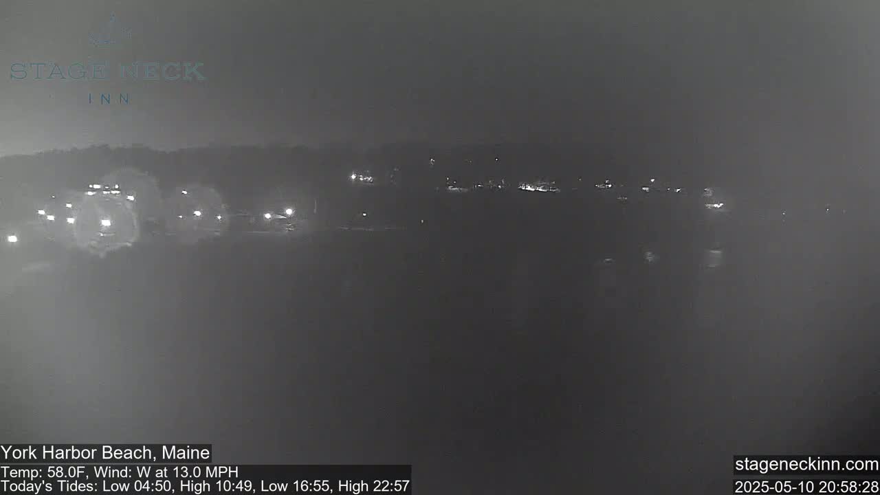 A nighttime, hazy view of a dark shoreline with faint lights visible in the distance, under a cloudy sky with a 13 mph west wind.