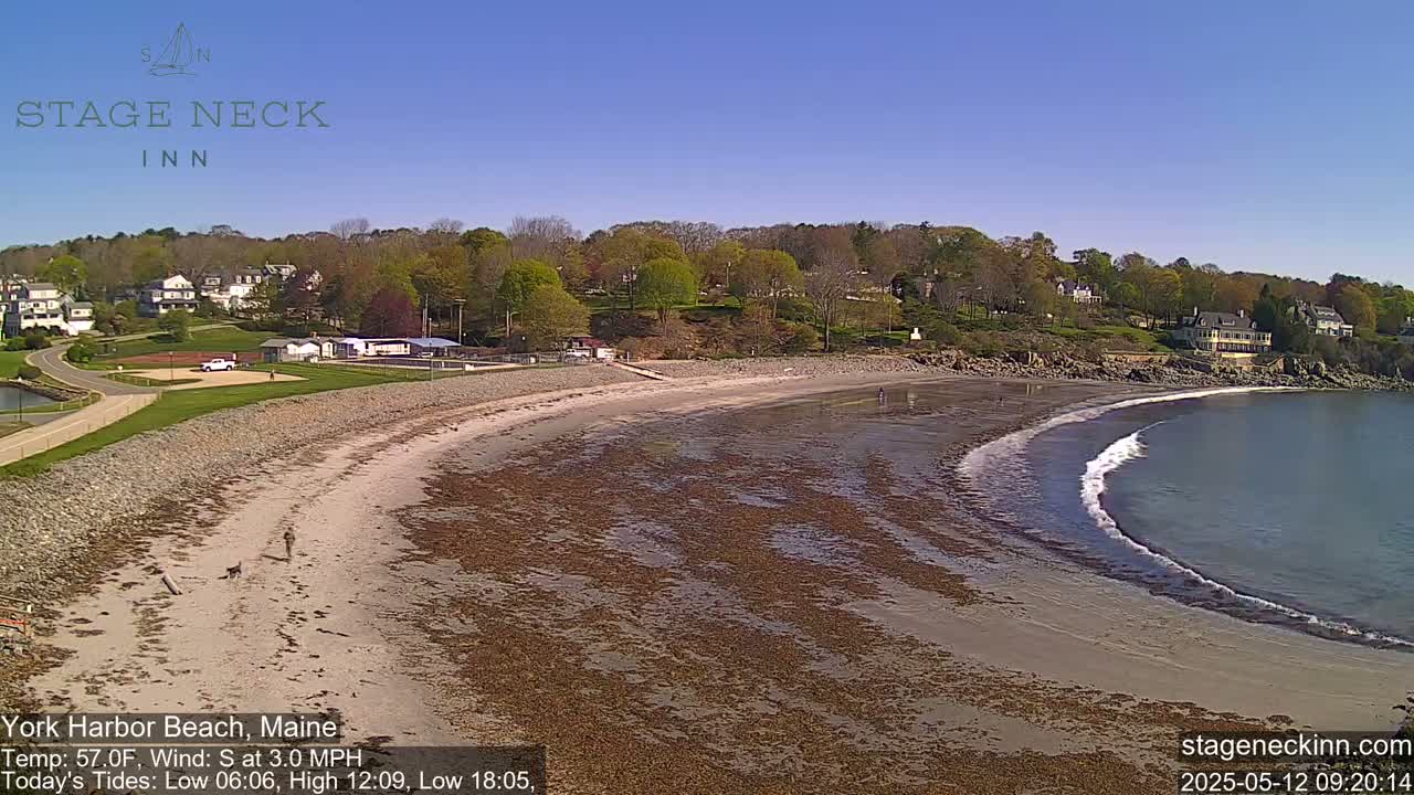 A sandy beach curves into a calm bay under a clear blue sky, with a few people walking along the shore and buildings visible in the background.