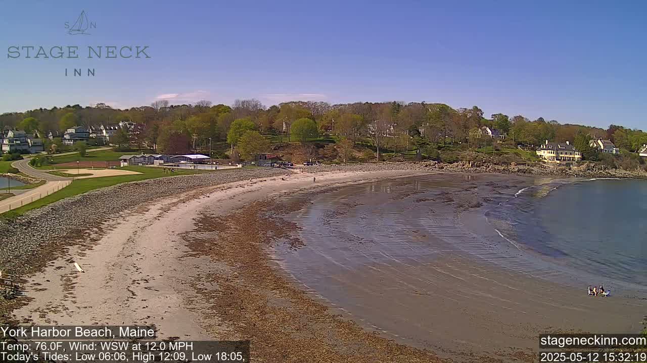 A sandy beach curves along a calm bay under a clear blue sky, with houses and trees visible on the hillside above.