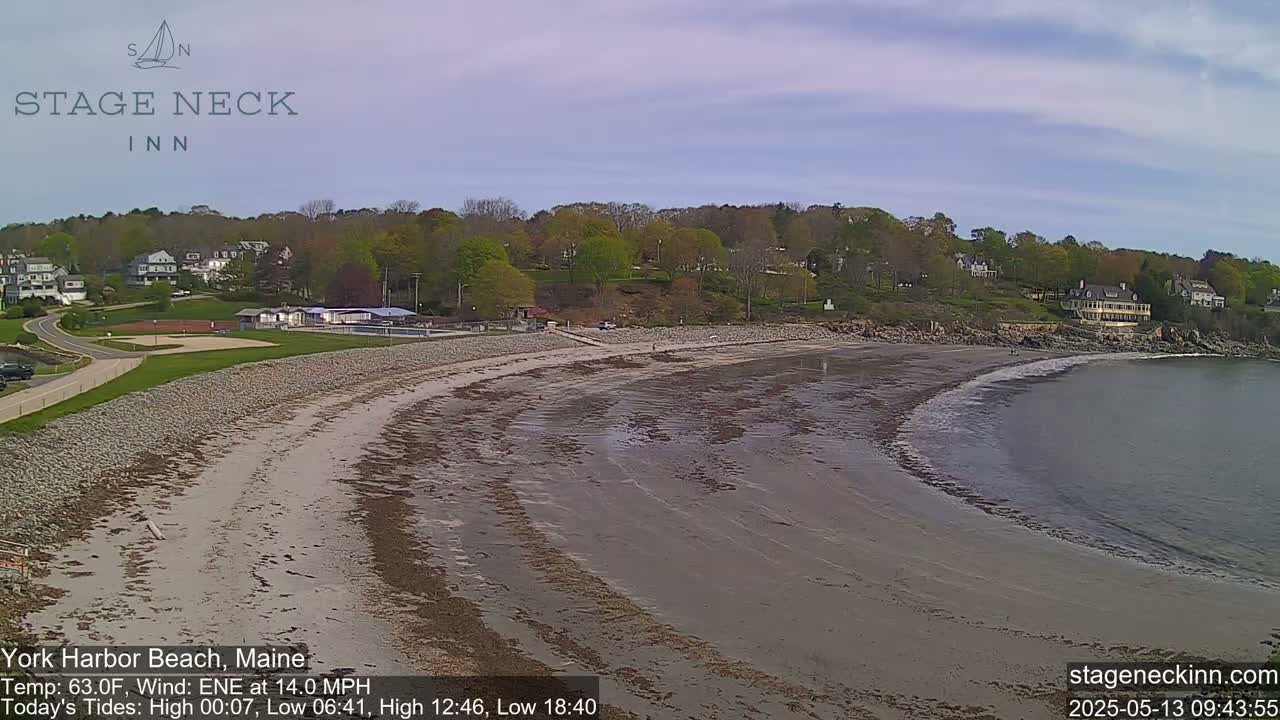 A sandy beach curves gently into a calm body of water under a partly cloudy sky, with green trees and buildings visible in the background.