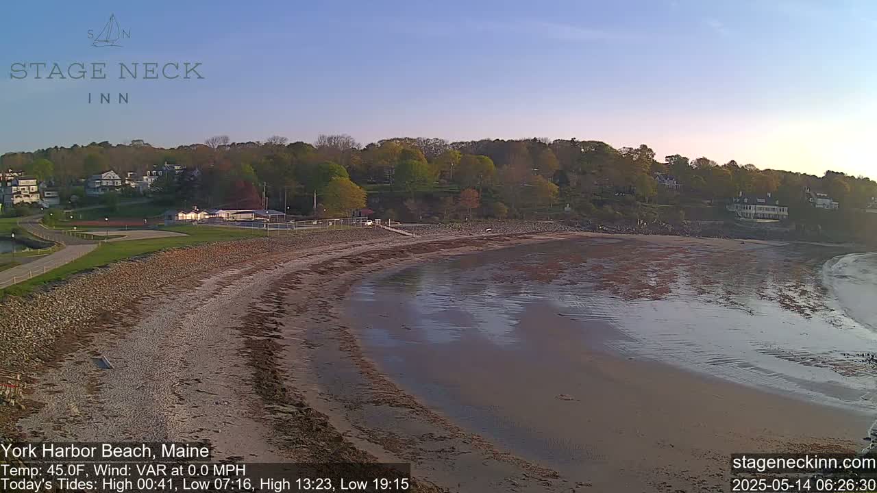 A sandy beach curves around a calm bay, backed by a line of trees and buildings under a clear, light-blue sky.