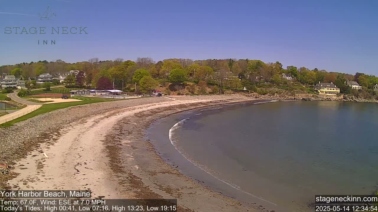 A sandy beach curves along a calm bay under a clear blue sky, bordered by lush green foliage and buildings.