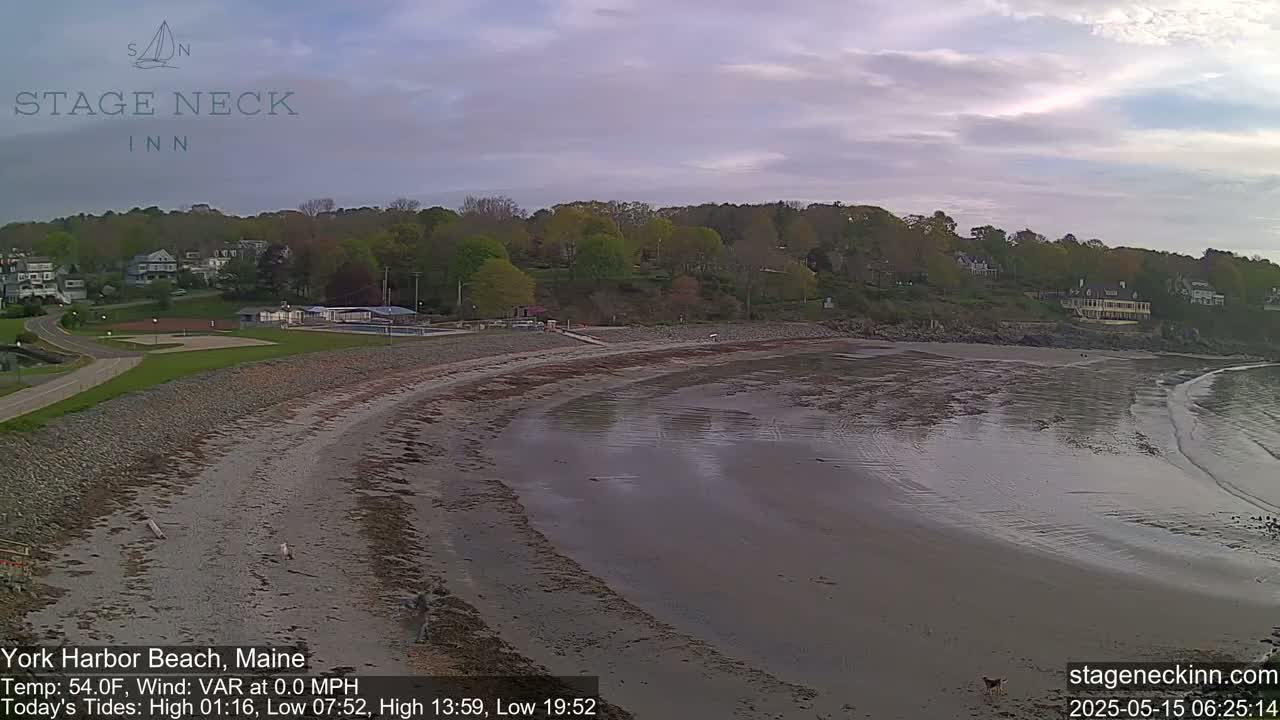 A mostly cloudy sky overlooks a sandy beach curving along a calm body of water, with green trees and buildings in the background.