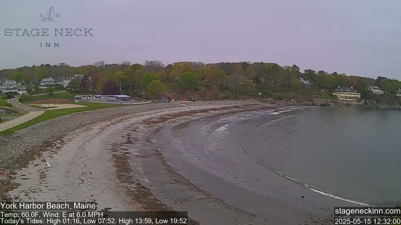 A sandy beach curves along a calm bay under an overcast sky, with green trees and buildings in the background.