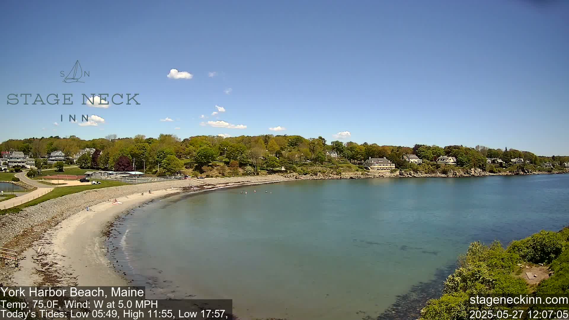 A sandy beach curves into a calm, teal bay under a mostly sunny sky, with houses and trees lining the shore.