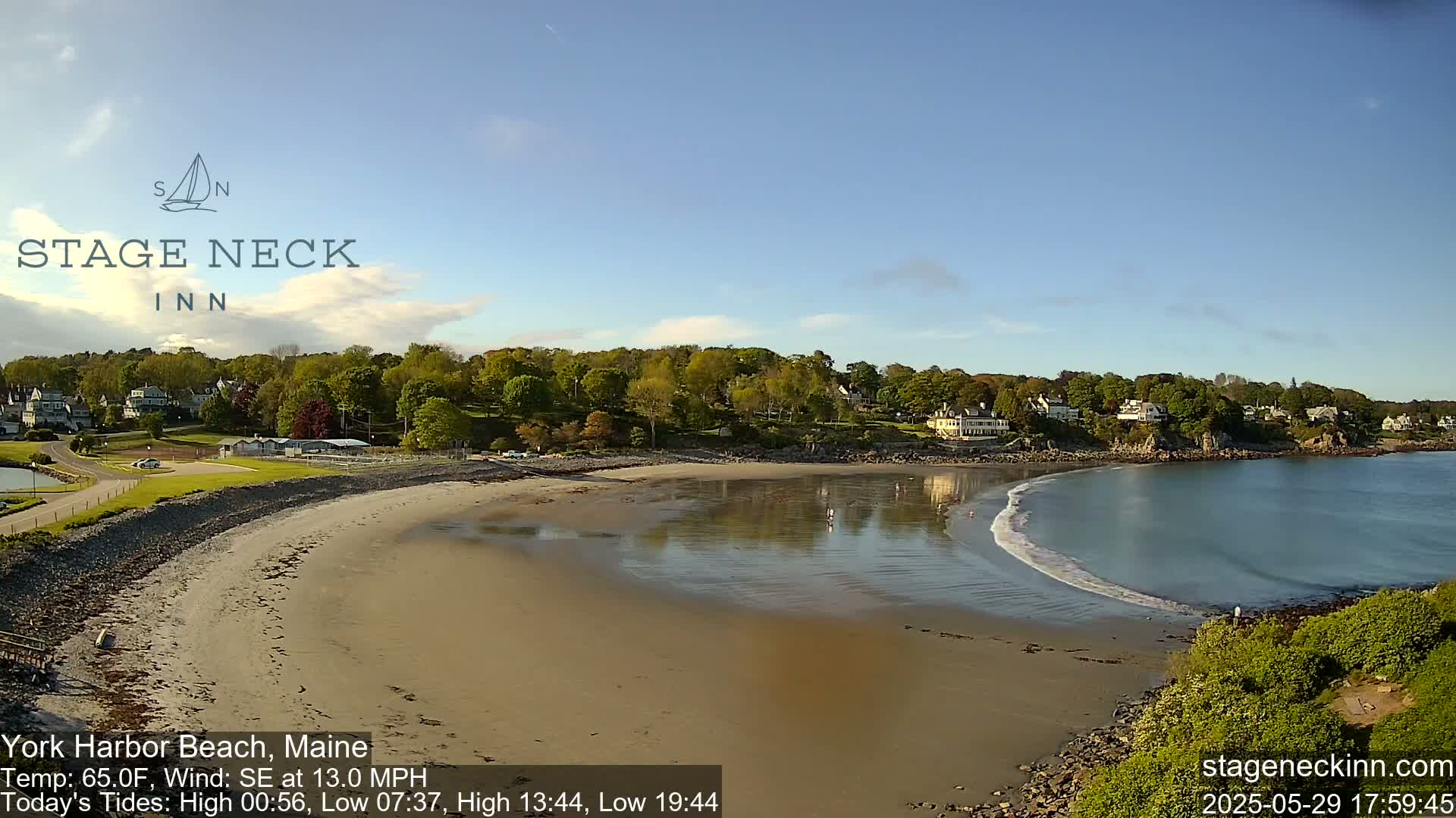 A sandy beach curves into a calm bay under a clear blue sky, with houses and trees lining the shore.