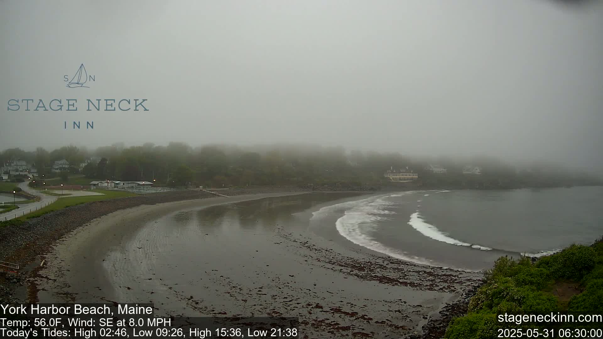 A foggy beach scene shows a sandy shore curving into a calm bay, with a small amount of wave action visible, and buildings and trees faintly visible in the distance.