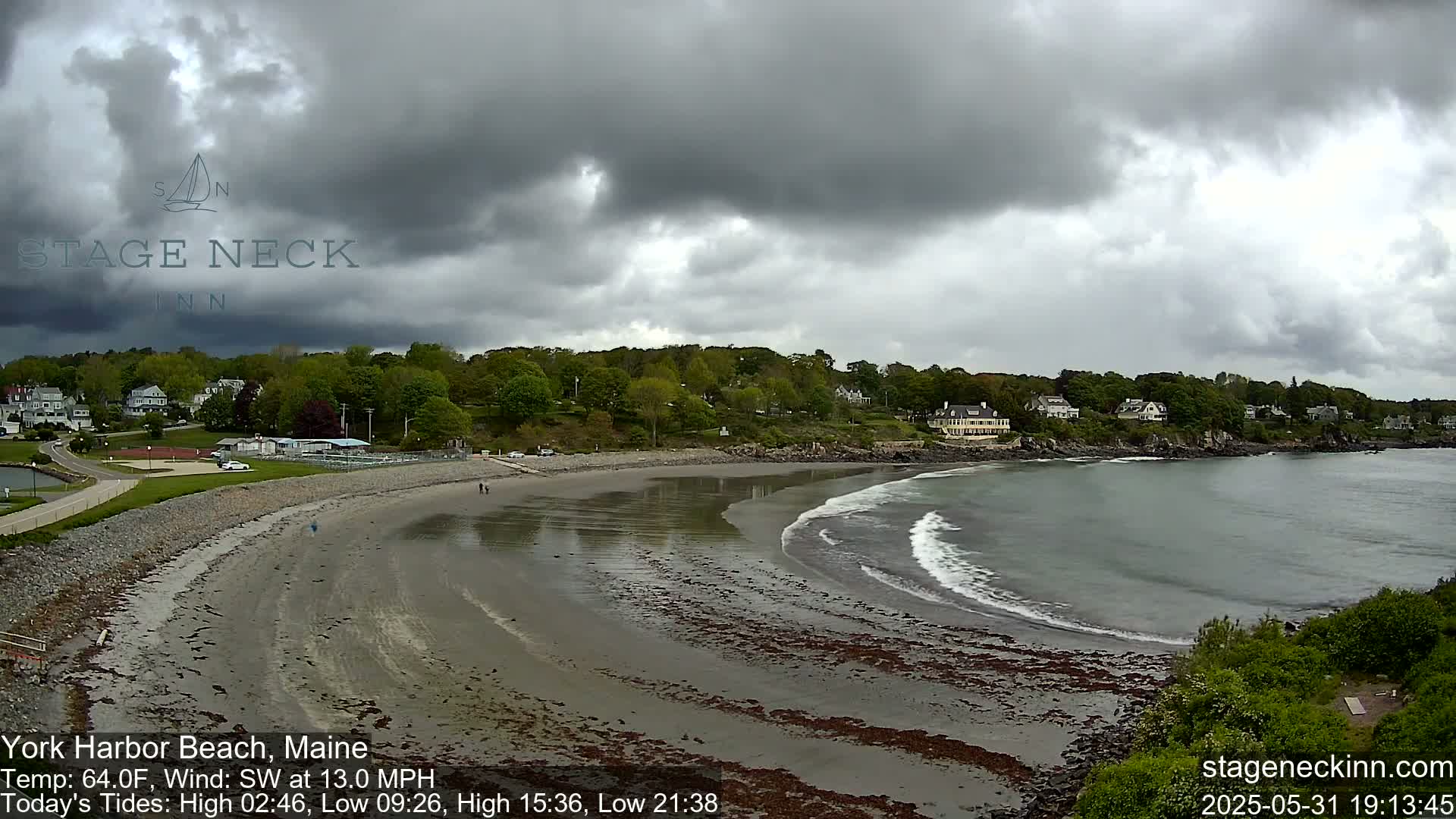 A sandy beach curves into a calm bay under a cloudy sky, with houses visible on a hillside in the background.