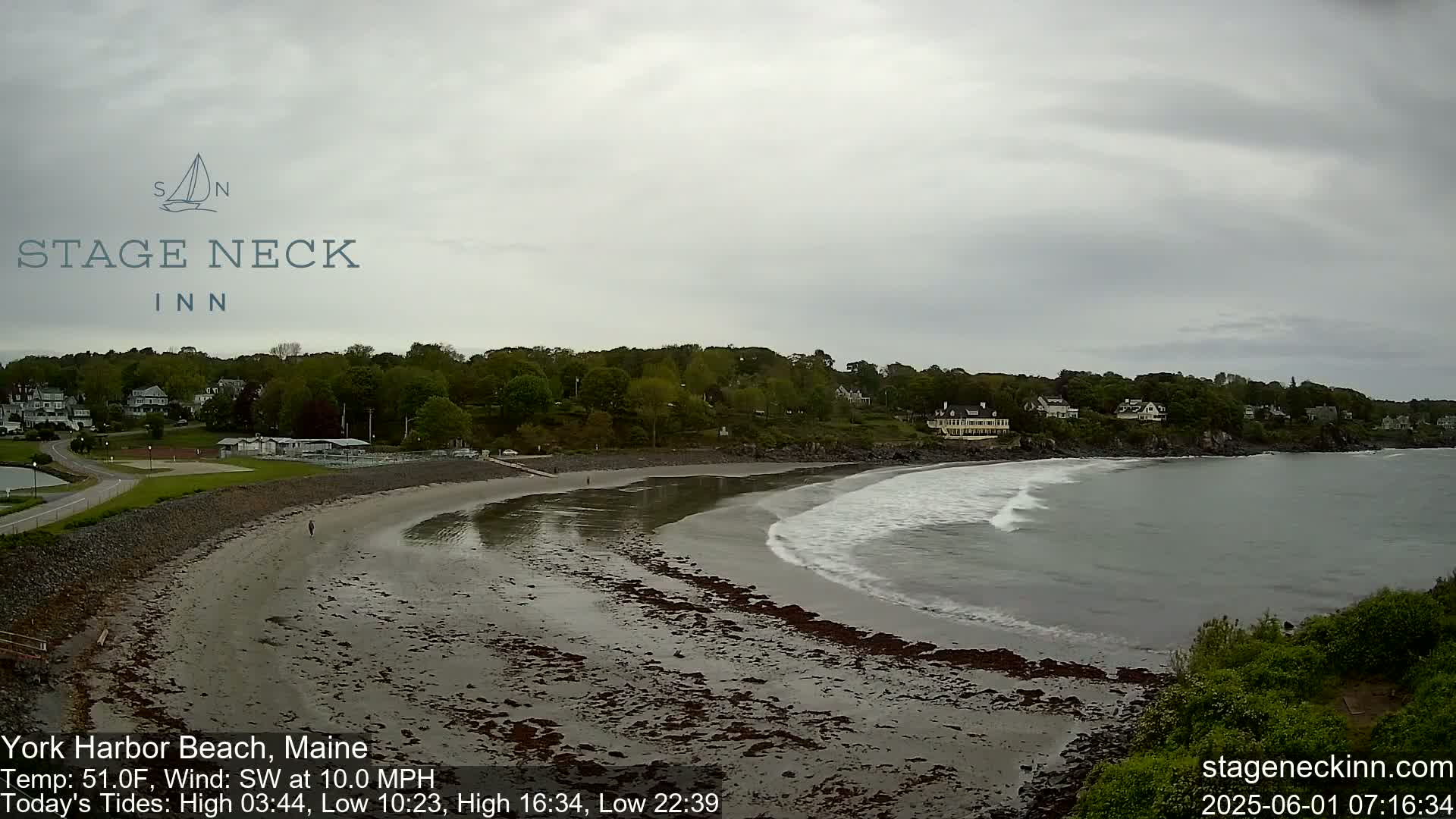 A sandy beach curves along a calm, gray ocean under an overcast sky, with houses and trees visible on the adjacent hillside.