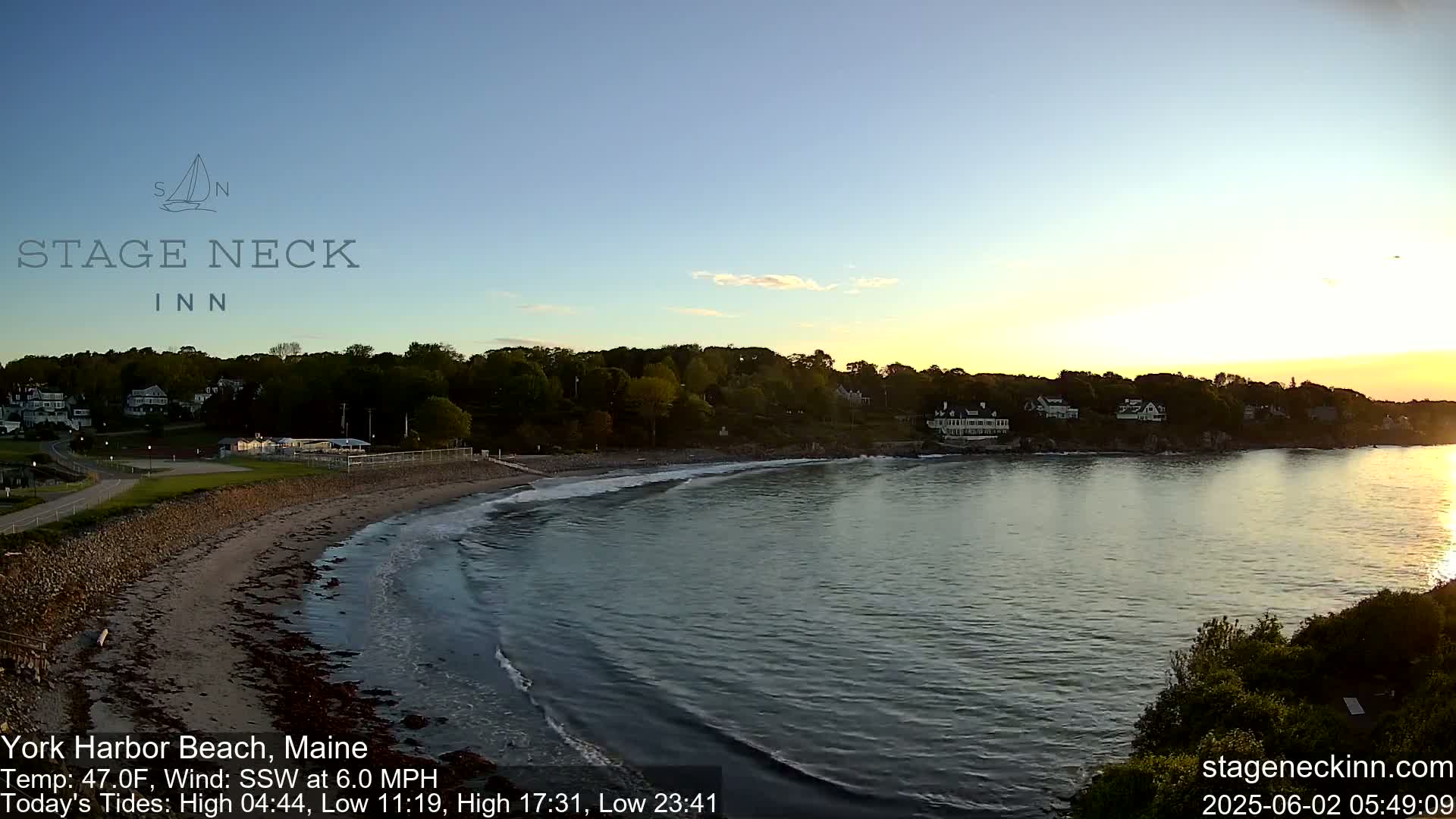 A sandy beach curves into a calm bay at sunset, with gentle waves lapping the shore and houses visible on the opposite bank under a clear sky.