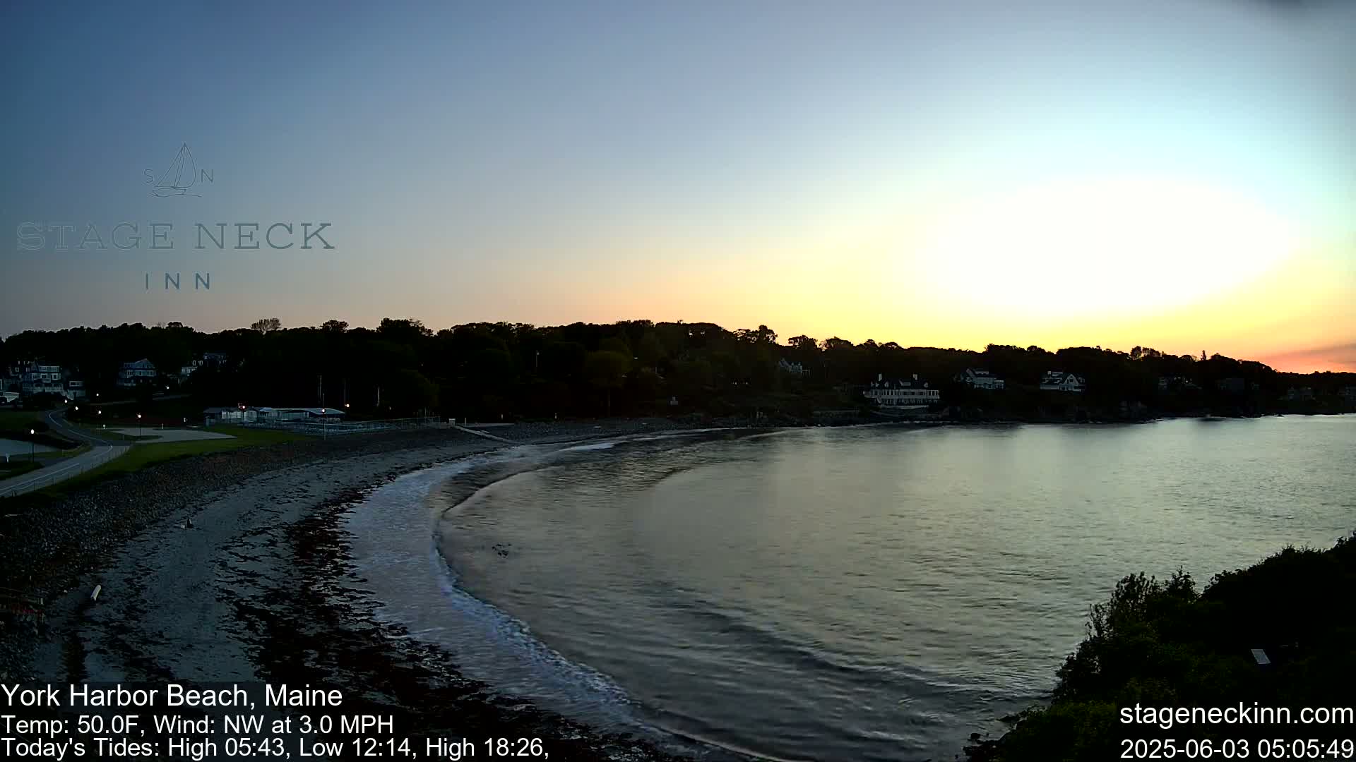 A calm bay at sunset with gentle waves lapping a dark sandy beach, backed by a tree line and houses.