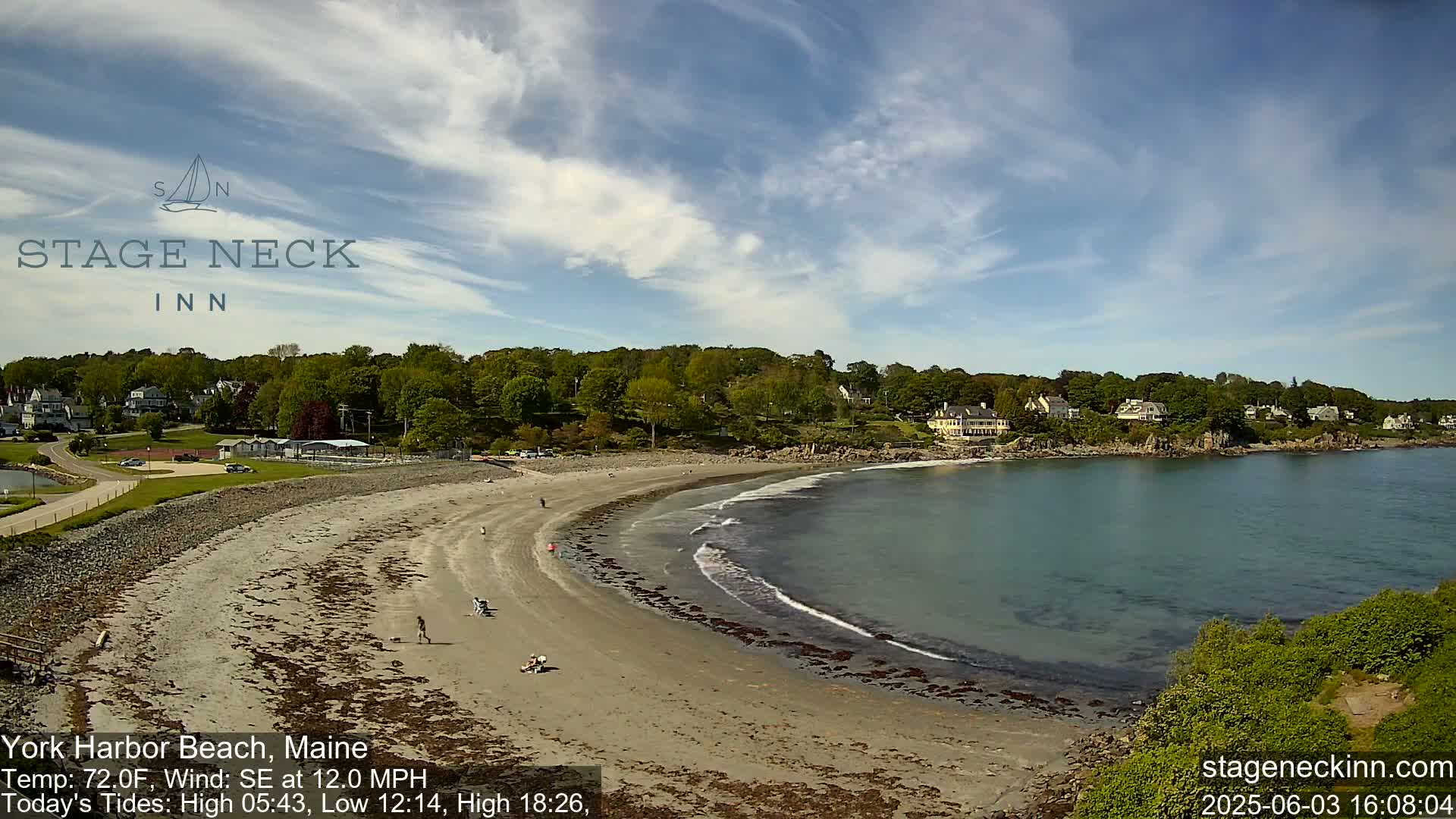 A sandy beach curves into calm ocean water under a partly cloudy sky, with houses and greenery visible on the hills beyond.