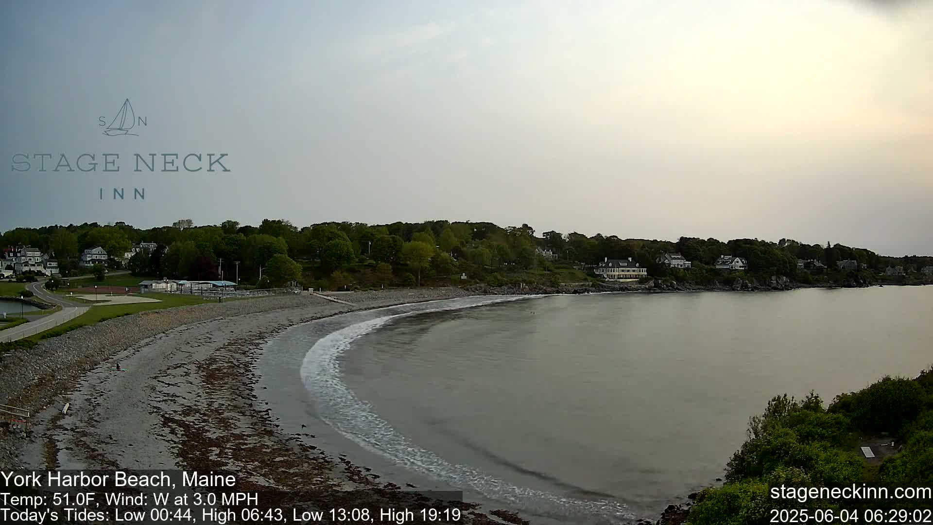 A mostly cloudy day shows a sandy beach curving into a calm bay, with a line of houses visible across the water.