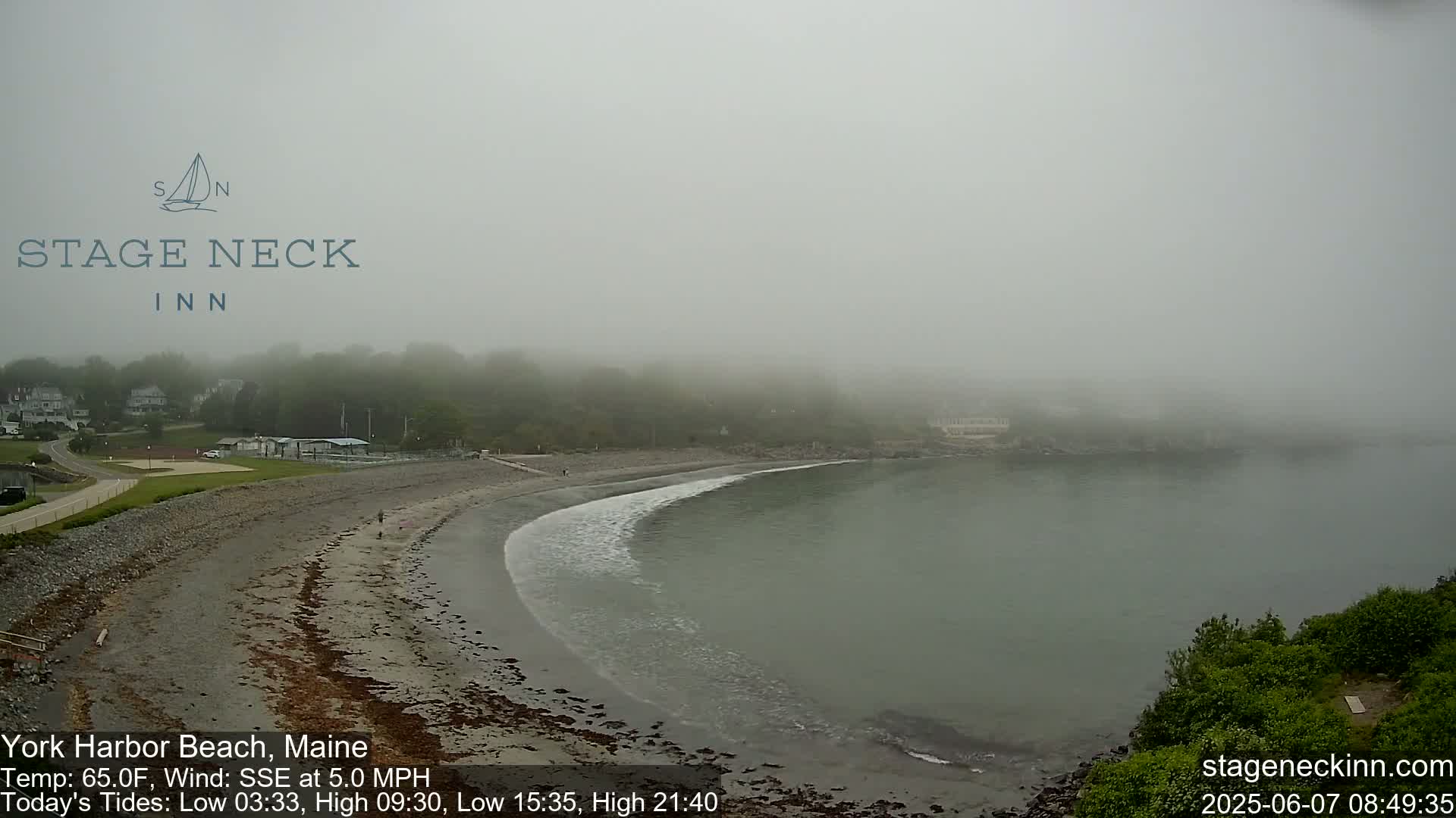 A foggy beach scene shows a calm bay, a sandy shore with a few people, and a line of trees and buildings shrouded in mist.