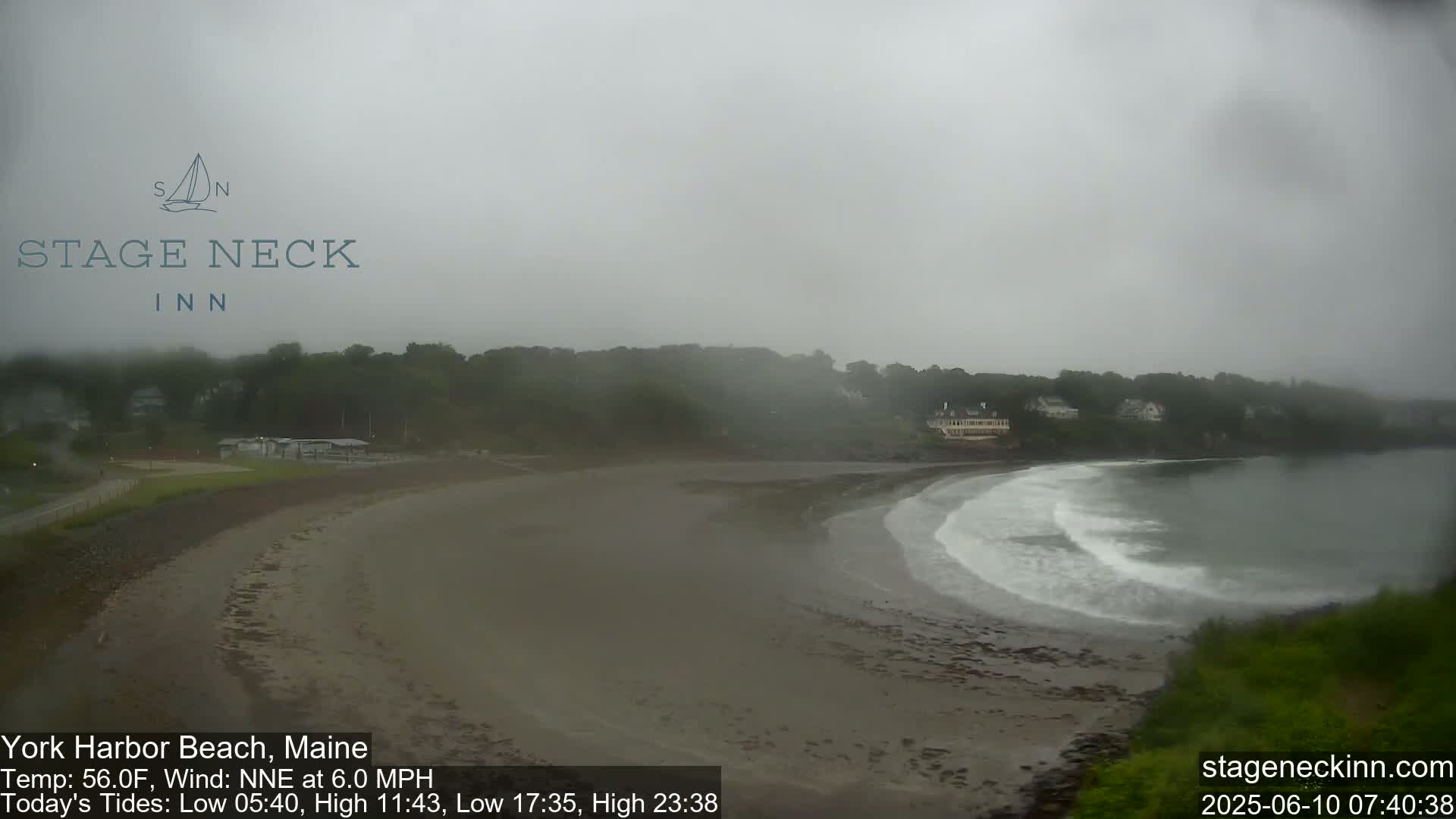 A foggy, overcast day at a sandy beach with gentle waves lapping the shore, and a line of trees and buildings visible in the distance.
