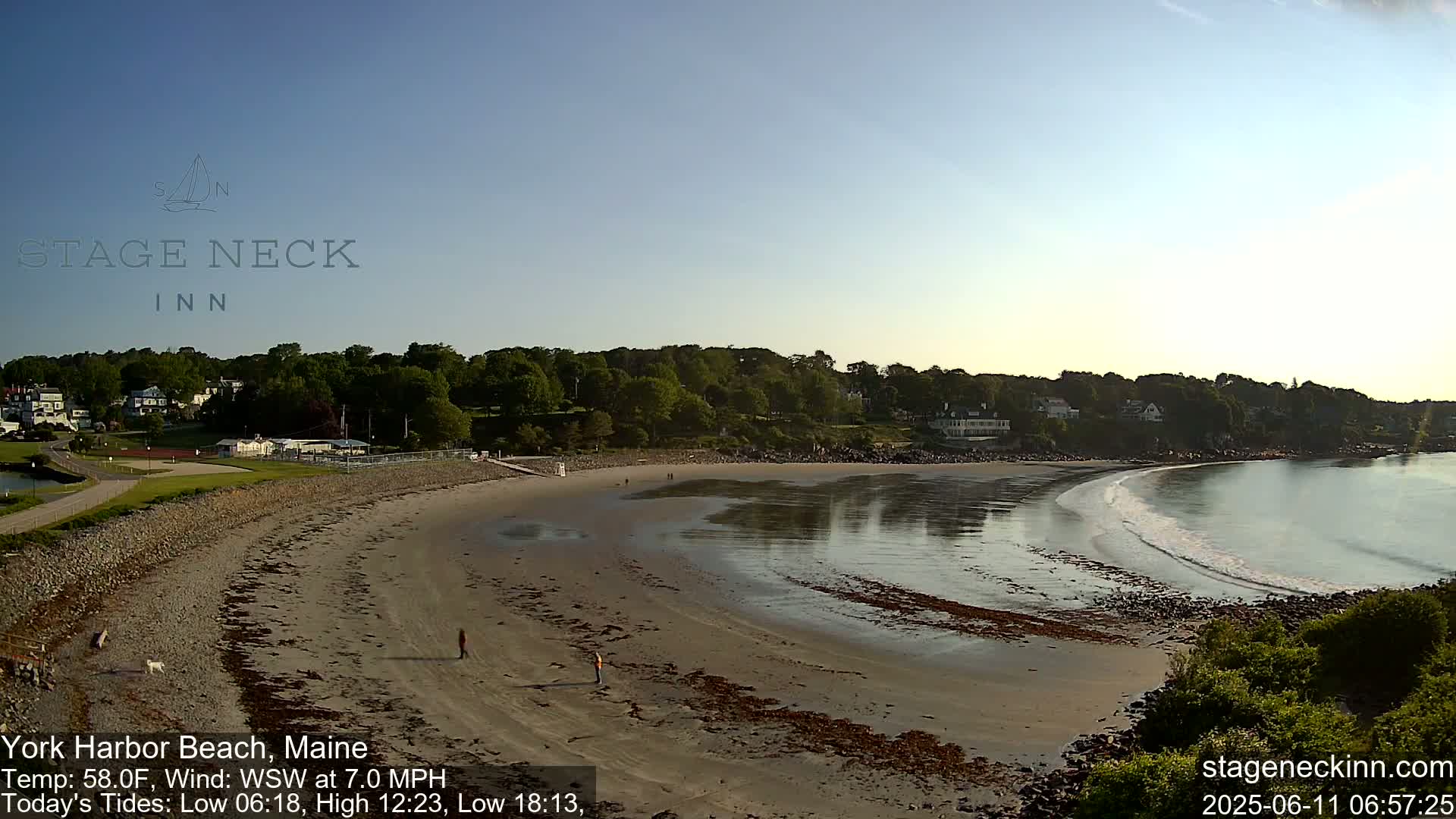 A sandy beach curves gently into calm, shallow water under a clear, bright sky, with a few people walking along the shore and buildings visible in the distance.