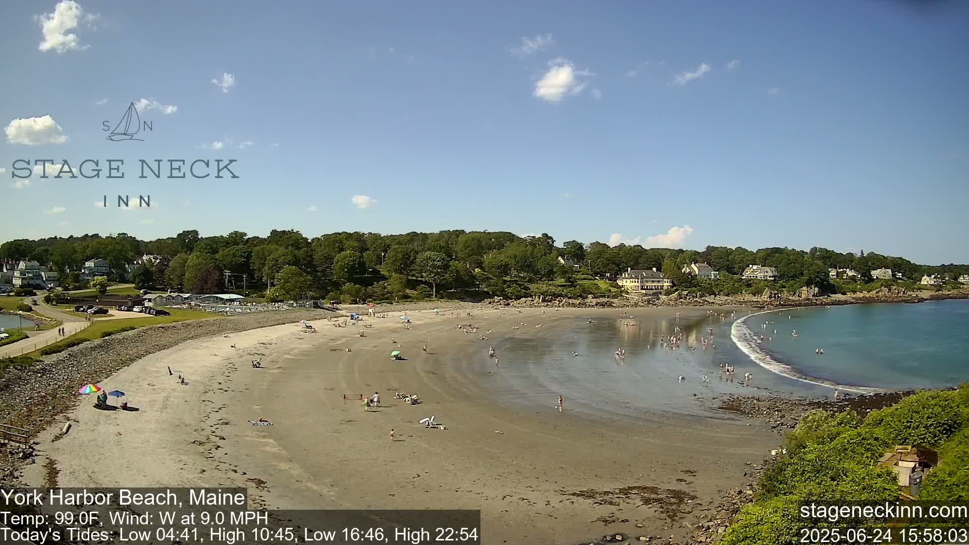 A sandy beach with many people swimming and relaxing, backed by green trees and houses, under a mostly sunny sky.
