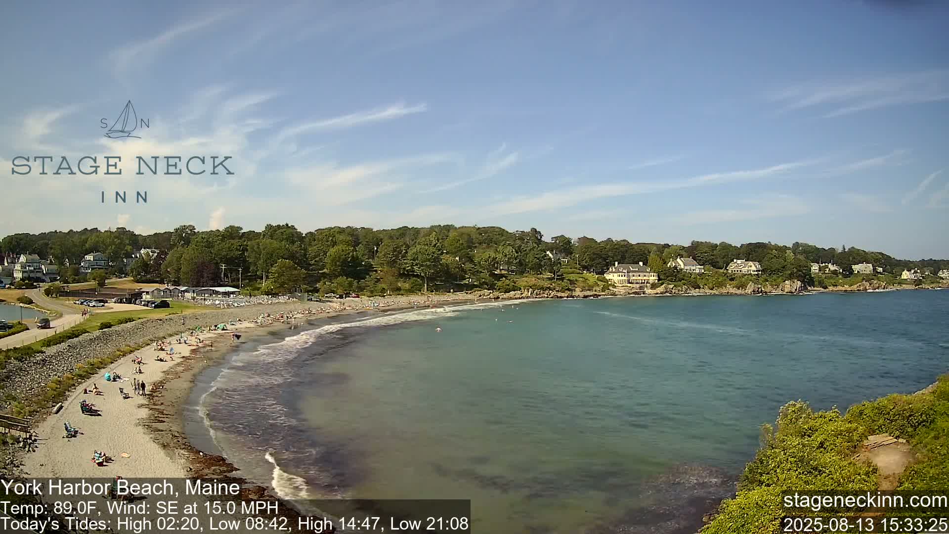 A sandy beach with people sunbathing and swimming is bordered by green trees and houses, under a mostly clear sky with a 15 mph southeast wind and 89°F temperature.