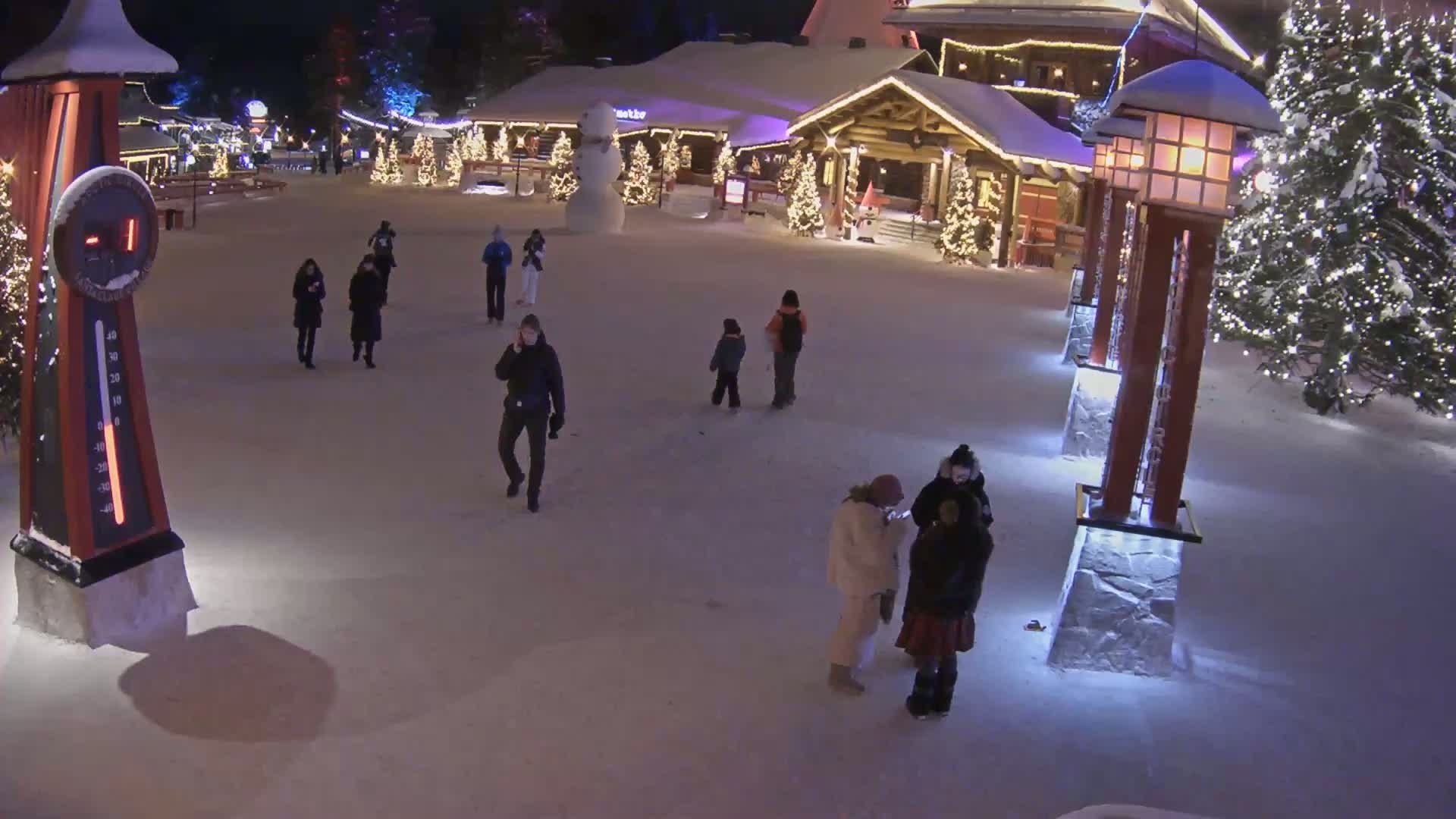 A snow-covered plaza at night, illuminated by lights on buildings and lampposts, features several people walking around and a snowman near a building.