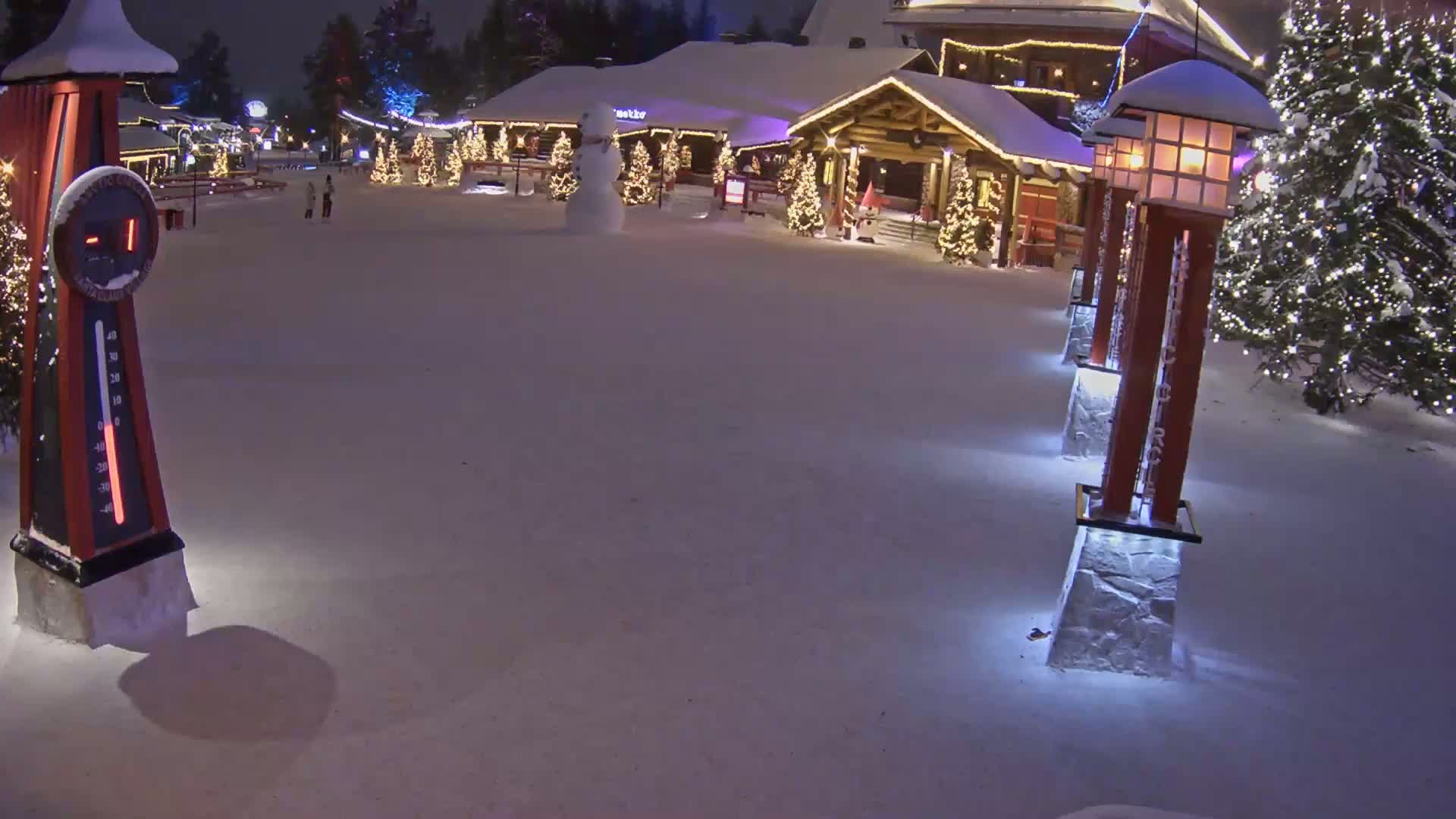 A snow-covered village square at night features a snowman, Christmas trees with lights, and several illuminated lampposts, with a thermometer indicating a temperature below zero.