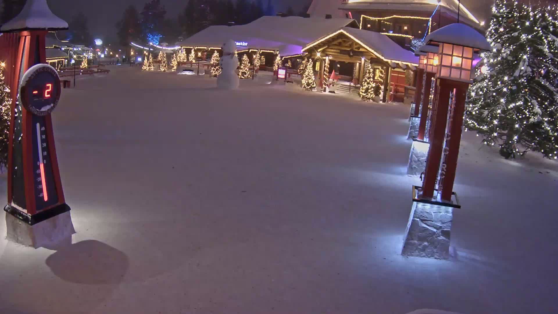 A snow-covered plaza at night, illuminated by lights on several posts and Christmas trees, shows a thermometer reading -2 degrees, a snowman, and a building decorated for the holidays.