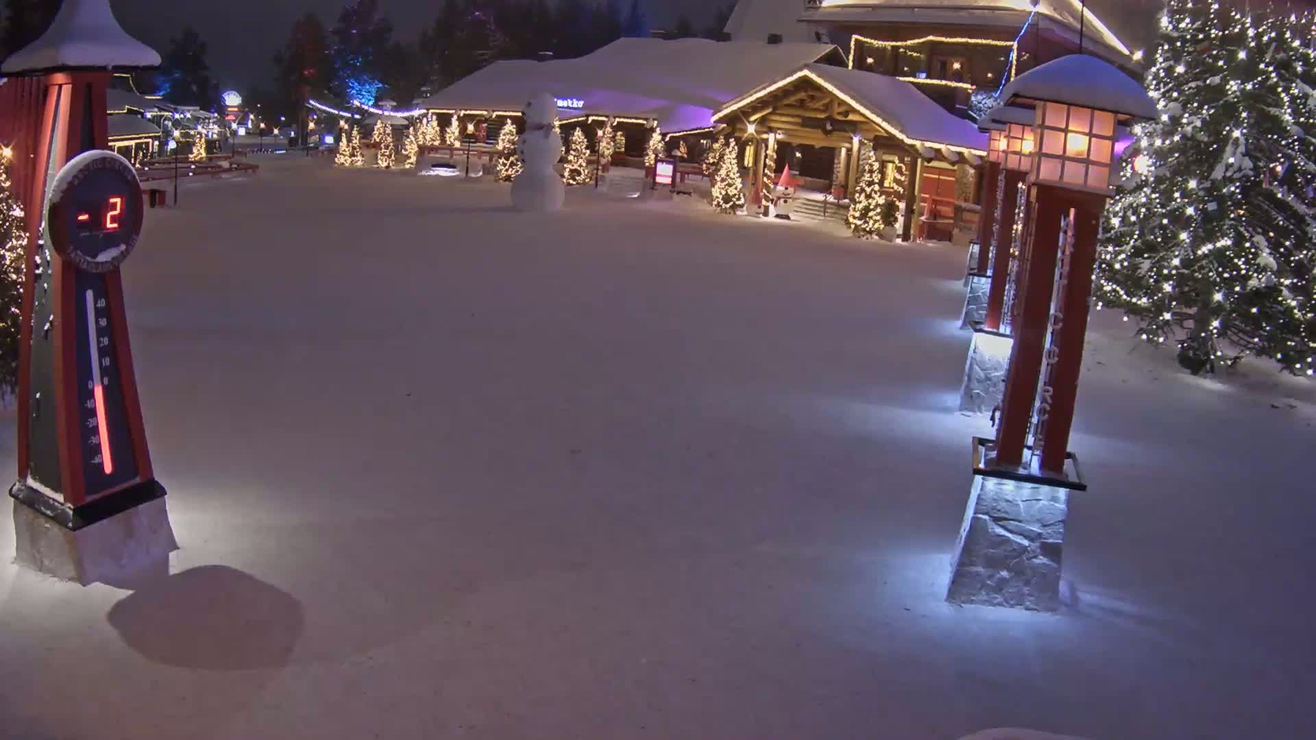 A snow-covered plaza at night, illuminated by lights on buildings and lampposts, shows a snowman and a thermometer indicating -2 degrees.