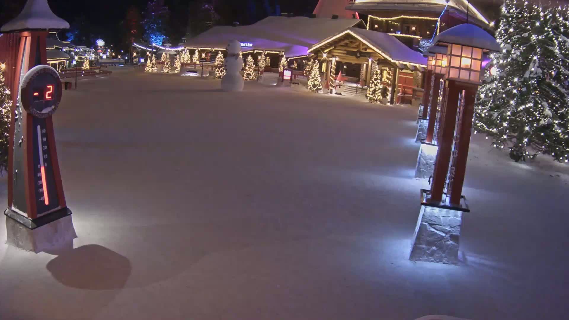 A snow-covered plaza at night is illuminated by lights on lampposts and Christmas trees, with a snowman standing near a building and a thermometer showing a temperature of -2 degrees.