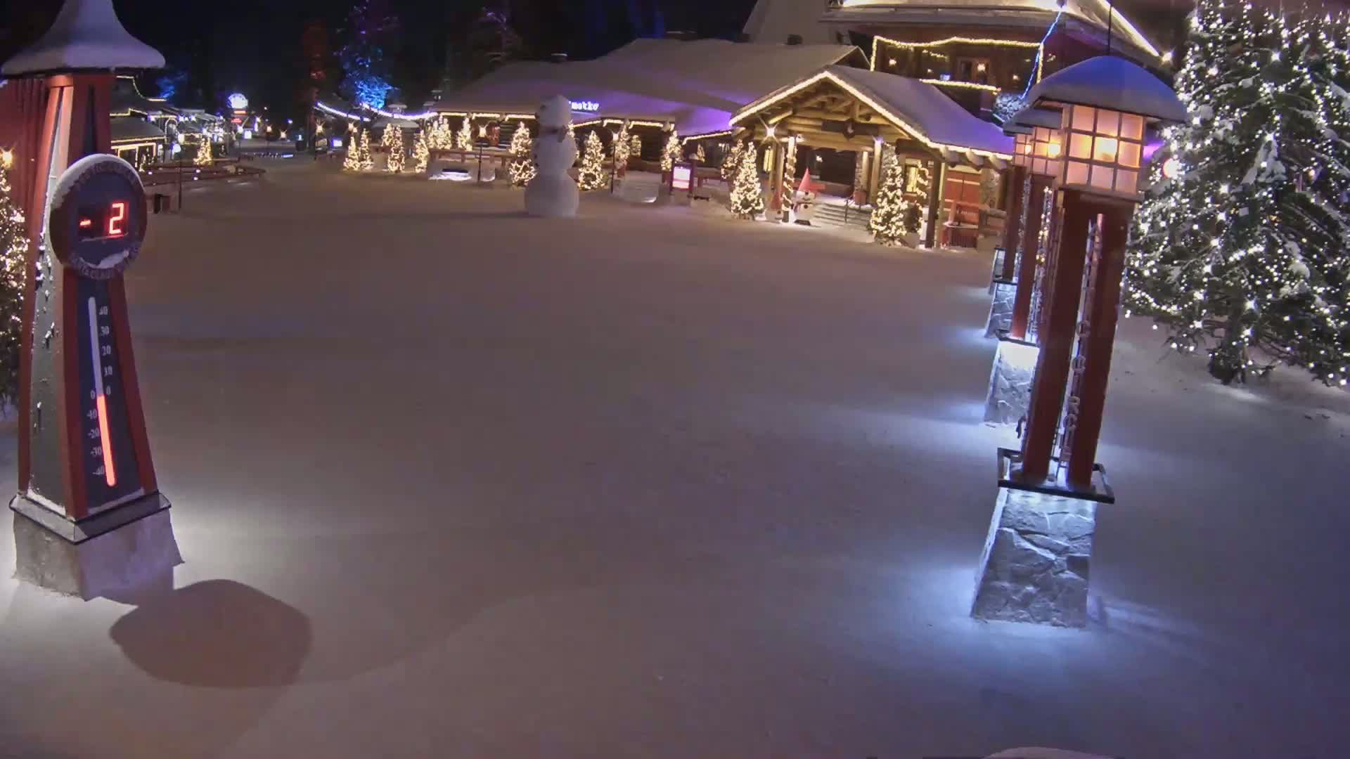 A snow-covered plaza at night, illuminated by lights on buildings and decorative lampposts, features a snowman and shows a temperature of -2 degrees.