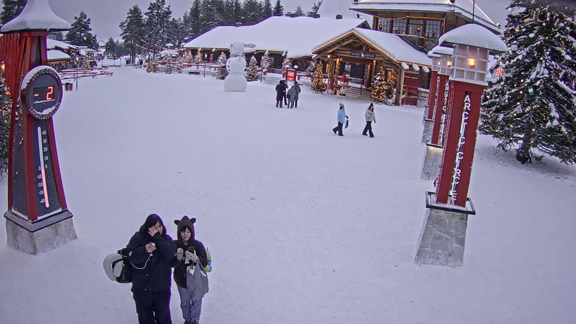 A snow-covered village scene shows several people walking past a snowman and a building, with two people standing close together in the foreground, in minus two-degree weather.