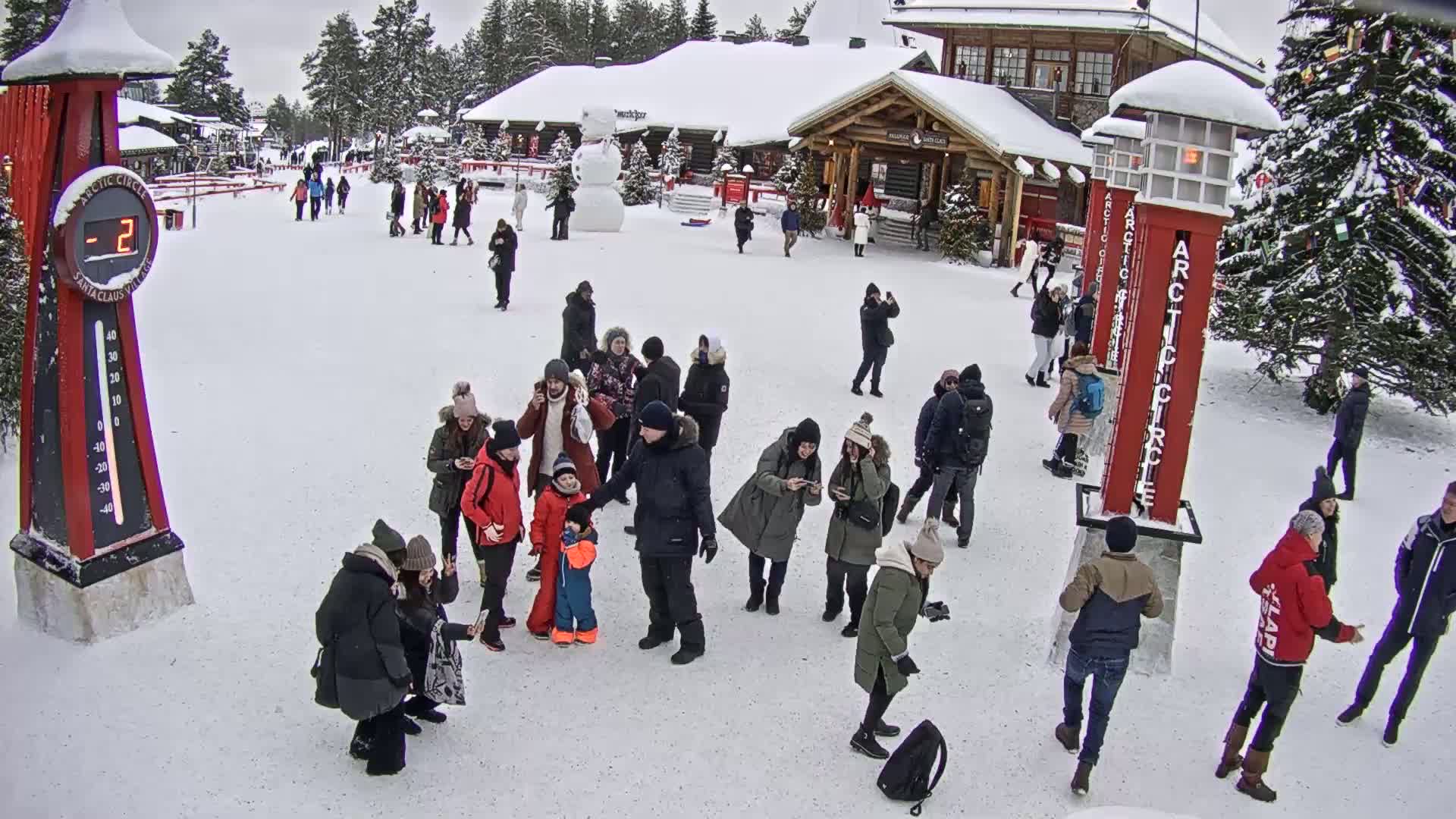 A snowy scene shows many people gathered in a plaza with wooden buildings in the background, a snowman, and a large thermometer displaying -2 degrees.