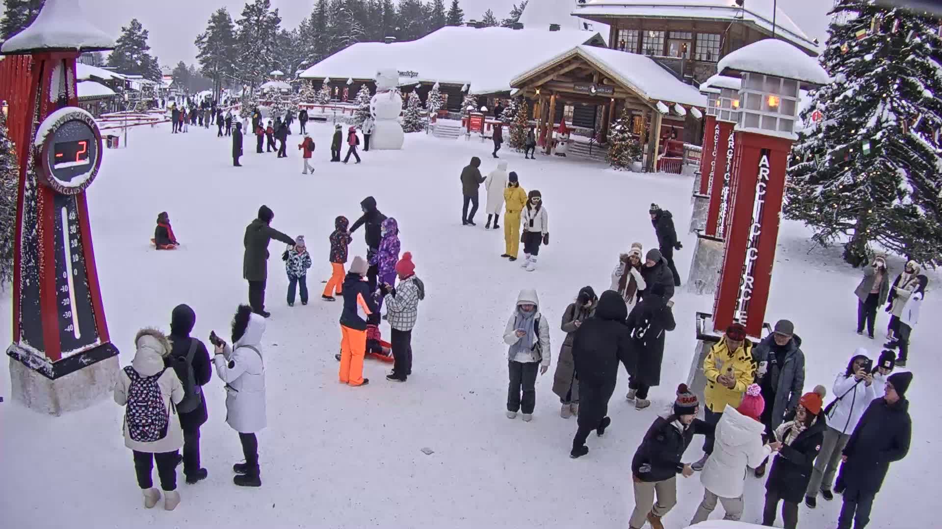 A snowy plaza with many people, some sitting and others standing in groups, near a building and a large snowman, under an overcast sky.
